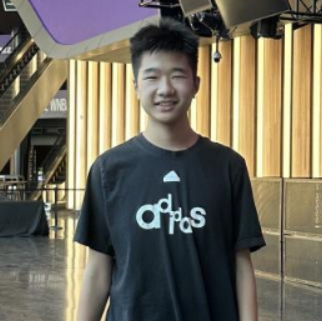 Young man smiling, wearing a black t-shirt with white text, standing indoors with wooden vertical panels in the background.