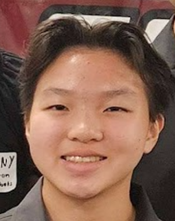 A young person with short dark hair smiling at the camera, wearing a gray top, in an indoor setting.