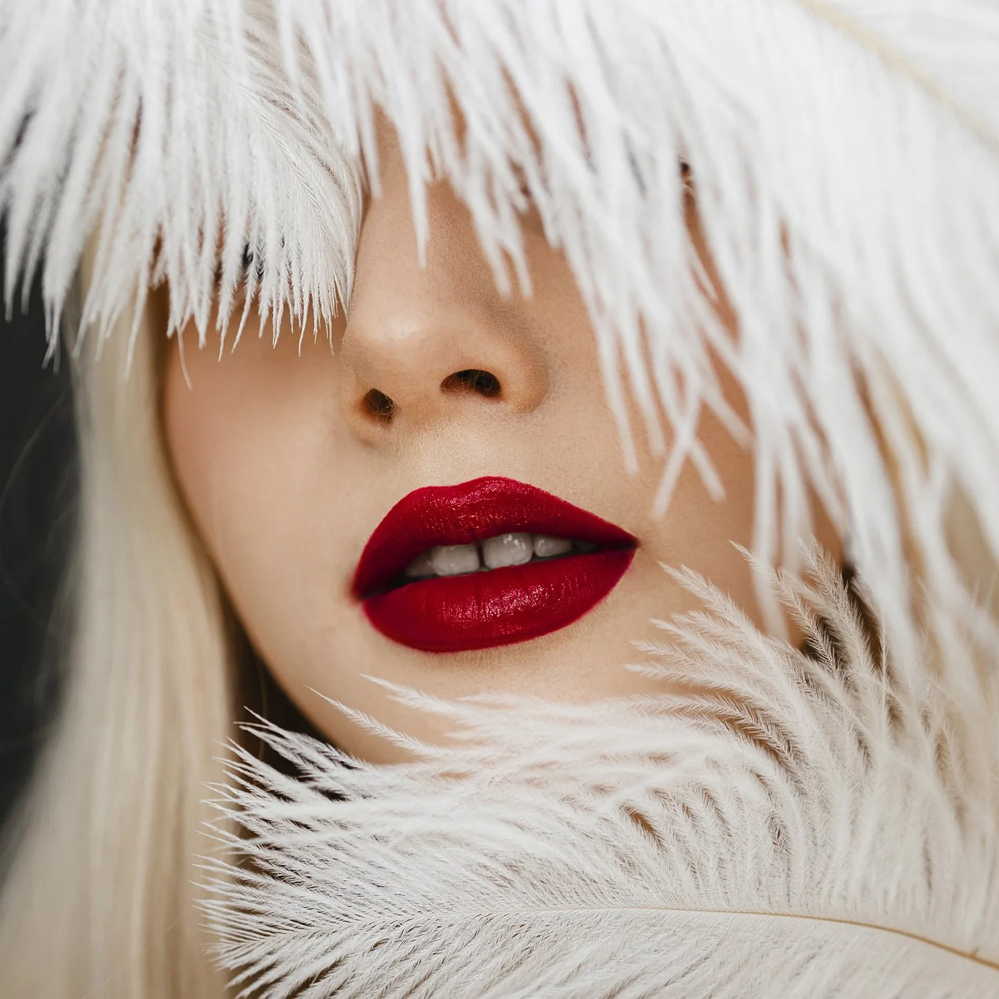 Close-up of a woman's face with platinum blonde hair, red lipstick, and partially covered eyes by white feathers.