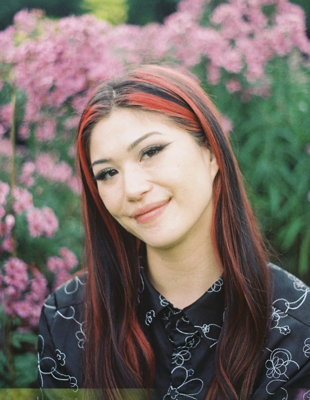 A young person with long, dark hair streaked with red, smiling outdoors in front of pink flowering bushes.