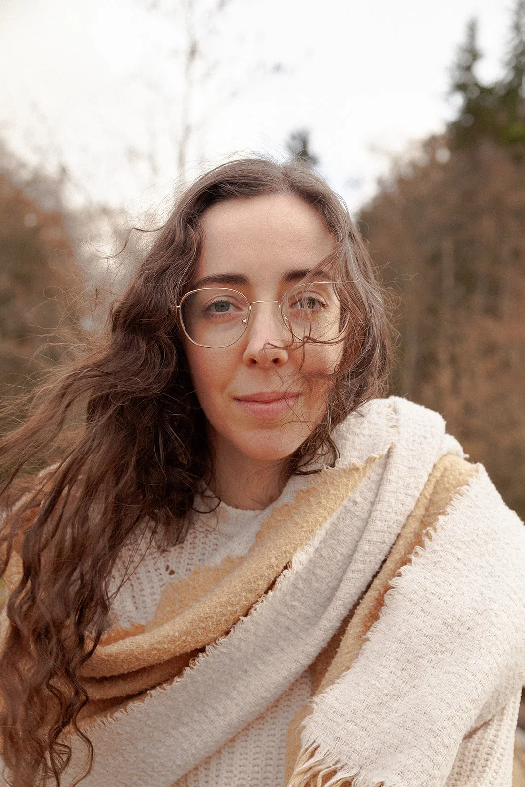 A young woman with curly brown hair and glasses, wearing a cream-colored, textured shawl outdoors amidst autumn trees.