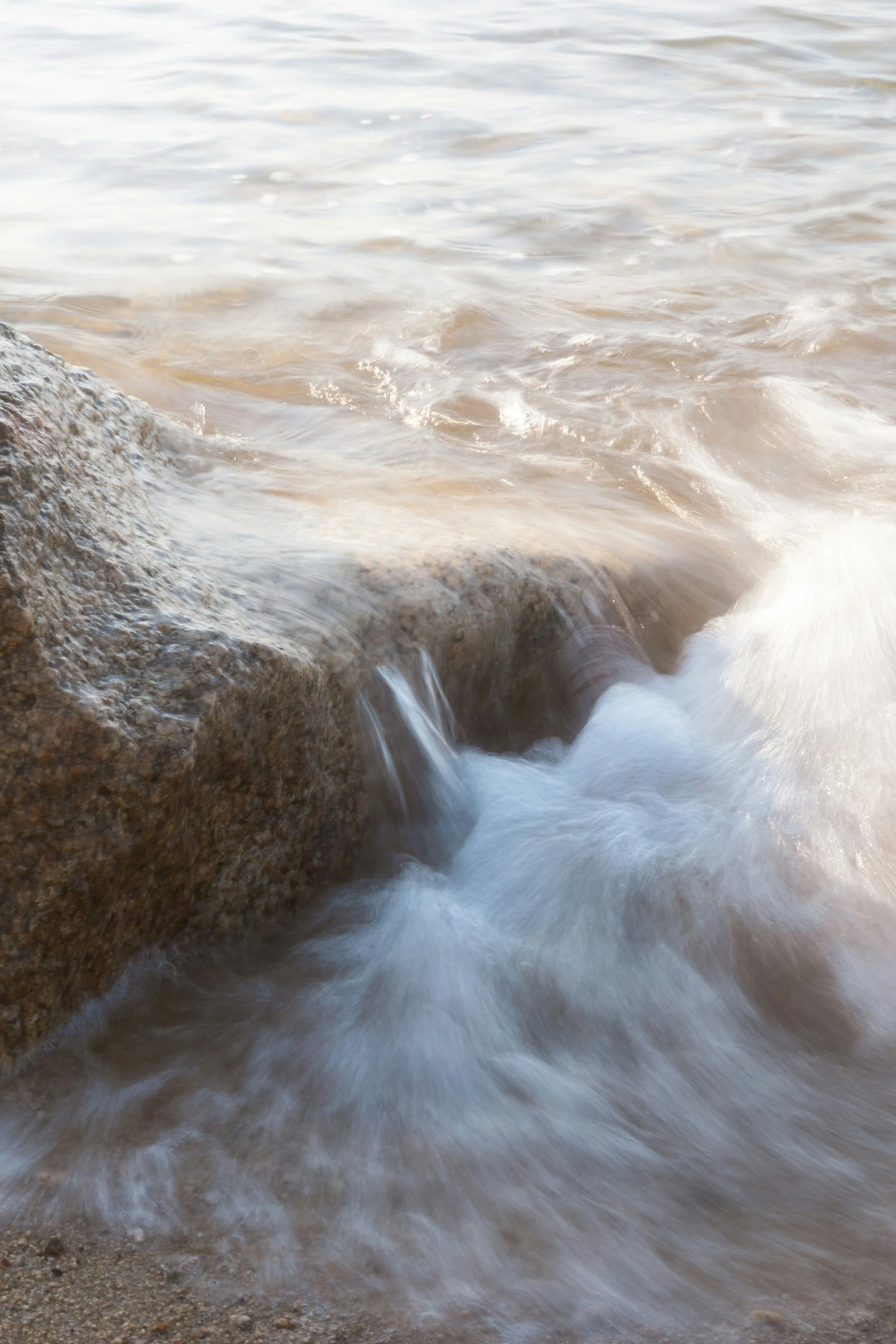 Waves crashing against rocks at the edge of a body of water, creating white foam.