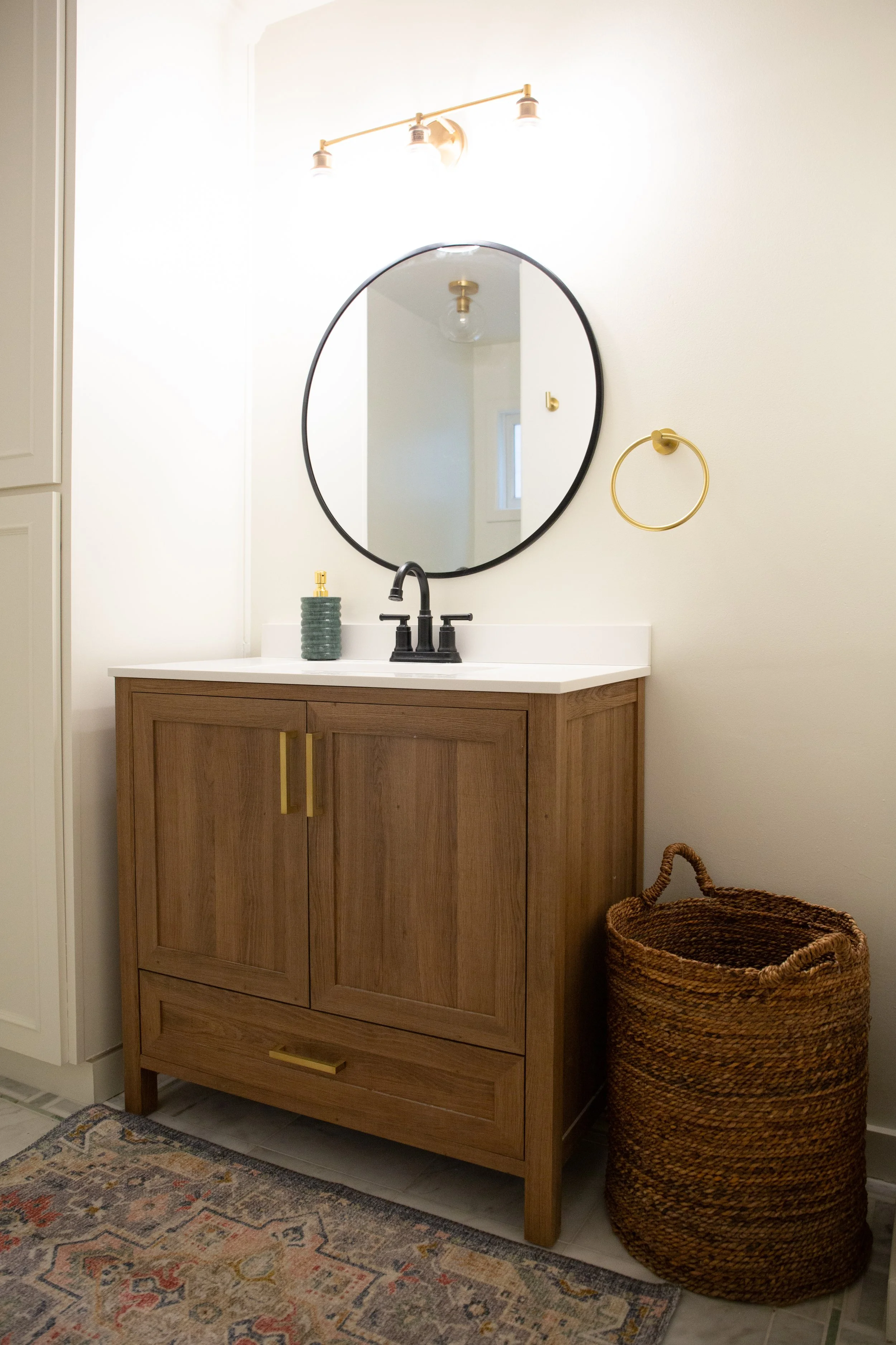 Bathroom vanity with a wooden cabinet, a round mirror, a black faucet, a soap dispenser, a gold towel ring, a basket, and a patterned rug.