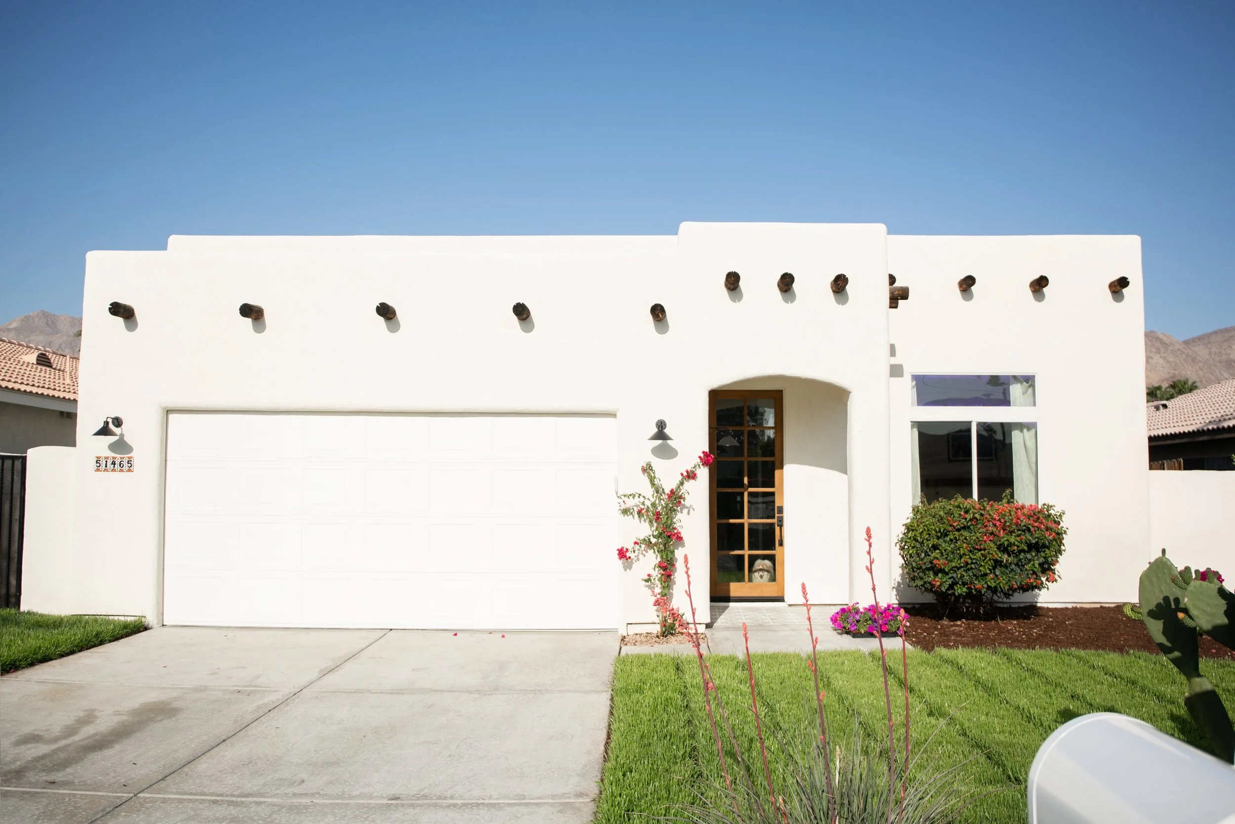 A modern white house with a flat roof, a garage door, a wooden front door with glass panels, a small window, and landscaped garden with bushes and flowers, under a clear blue sky.