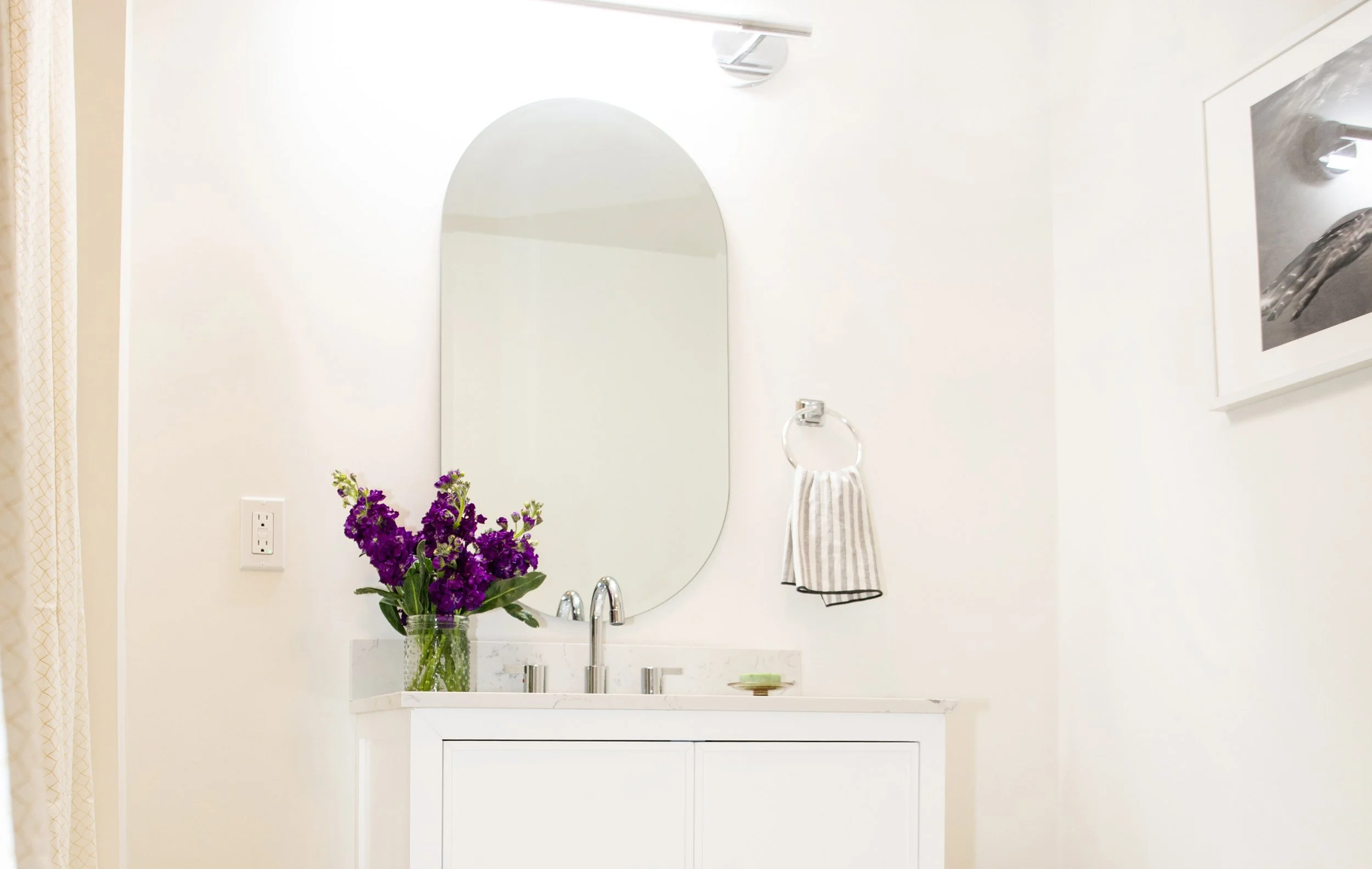 Bathroom vanity with a white cabinet, a marble countertop, a tall mirror, a vase of purple flowers, a soap inside a dish, a wall-mounted towel ring with a striped hand towel, and framed artwork on a white wall.
