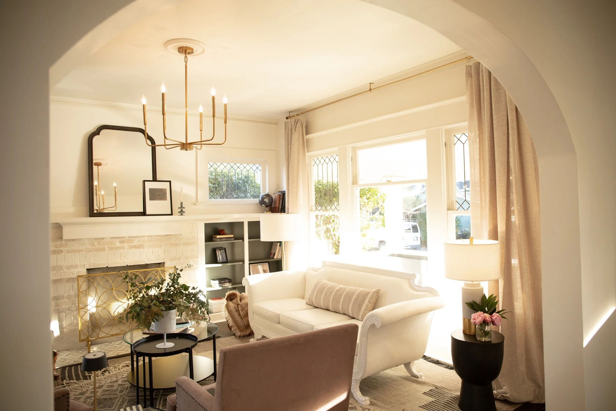 Bright living room with cream-colored sofa, pink armchair, black side table with pink flowers, white brick fireplace, built-in white shelves, gold chandelier, large windows with beige curtains, and decorative mirror and artwork.
