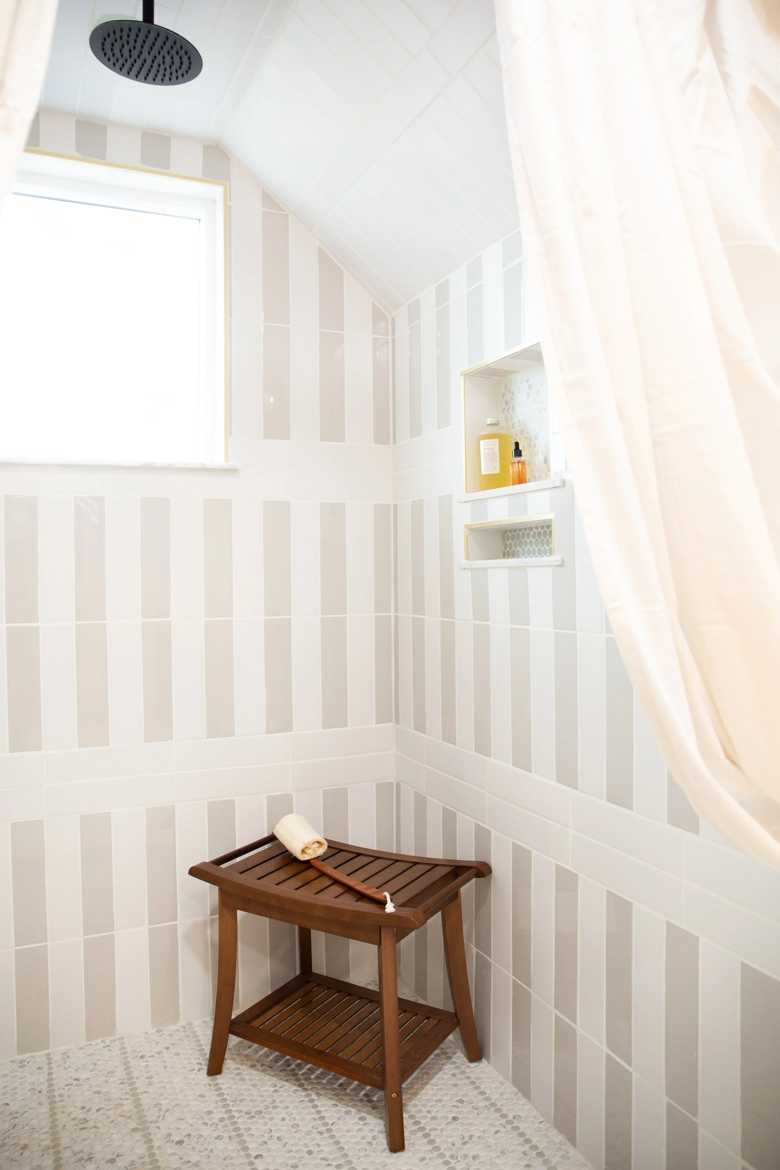 Bright, minimalist shower with white tiled walls, a ceiling rain shower, a small window, a wooden shower stool with a rolled towel, and built-in wall shelves with toiletries.