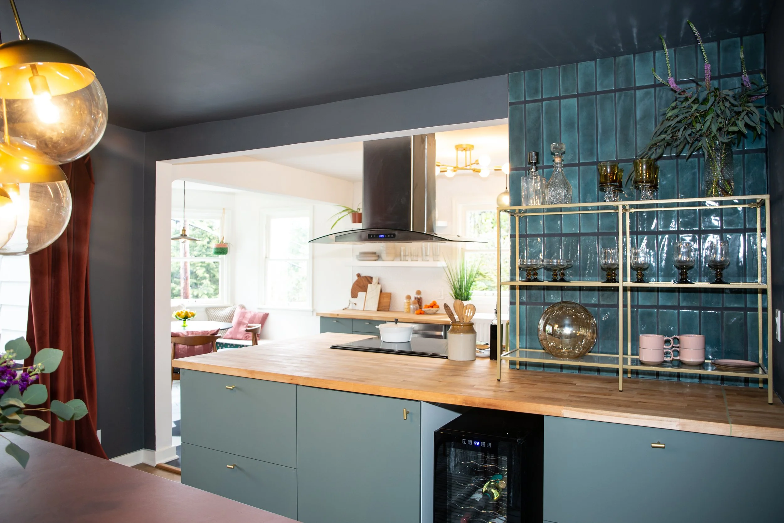 Kitchen with blue cabinets and a wooden countertop, blue tiled wall, glassware on a gold shelving unit, and a view into a bright dining area with windows, plants, and seating.