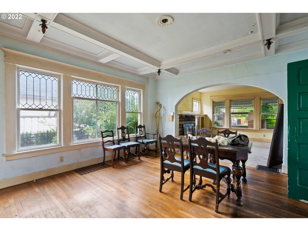 A dining room with wooden flooring and large windows with decorative grids, opening into a living room with a fireplace and lush green view outside.