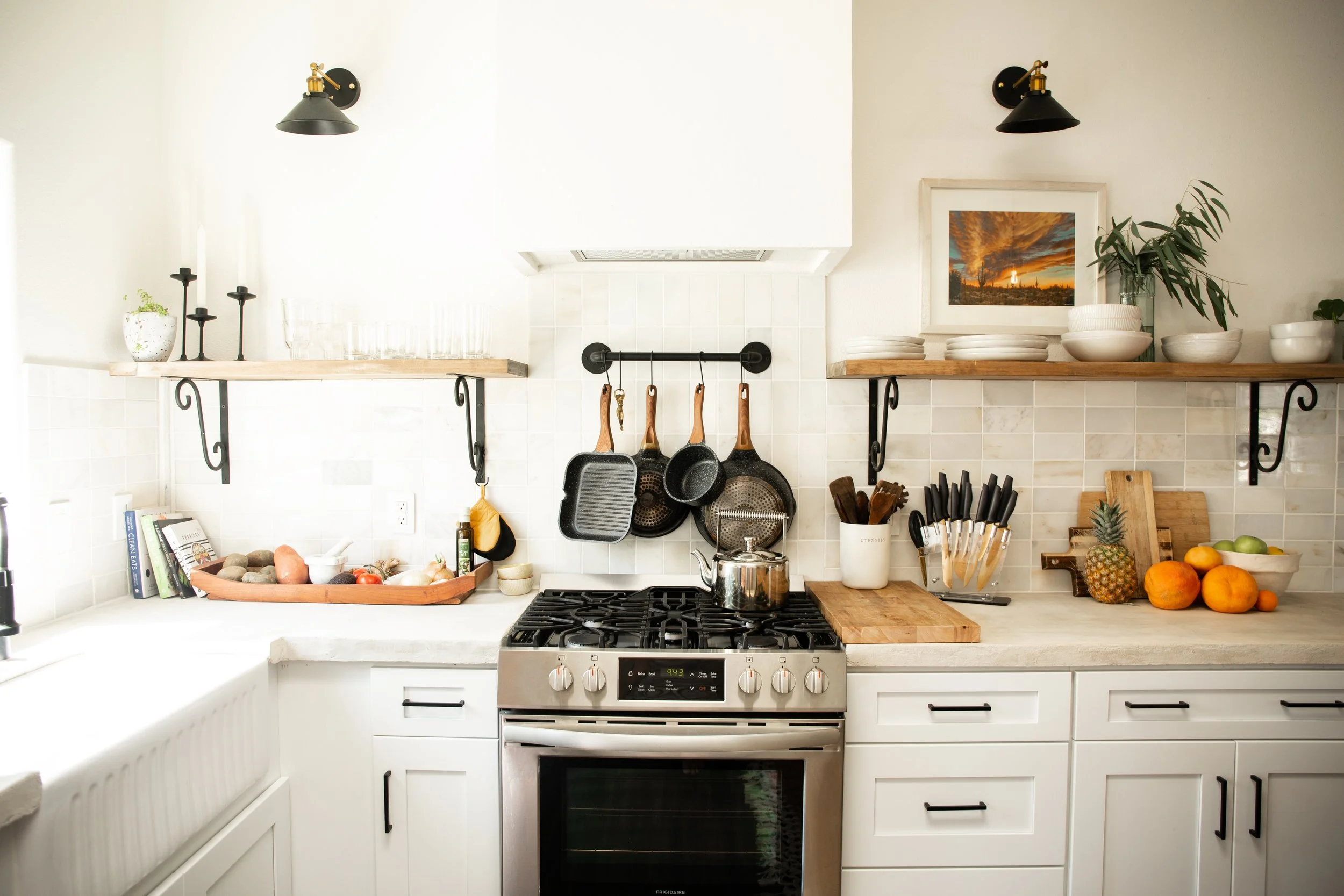 Modern kitchen with white cabinets, stainless steel oven, black wall sconces, wooden shelves with dishes and decorative items, hanging frying pans, and fresh fruit on the counter.