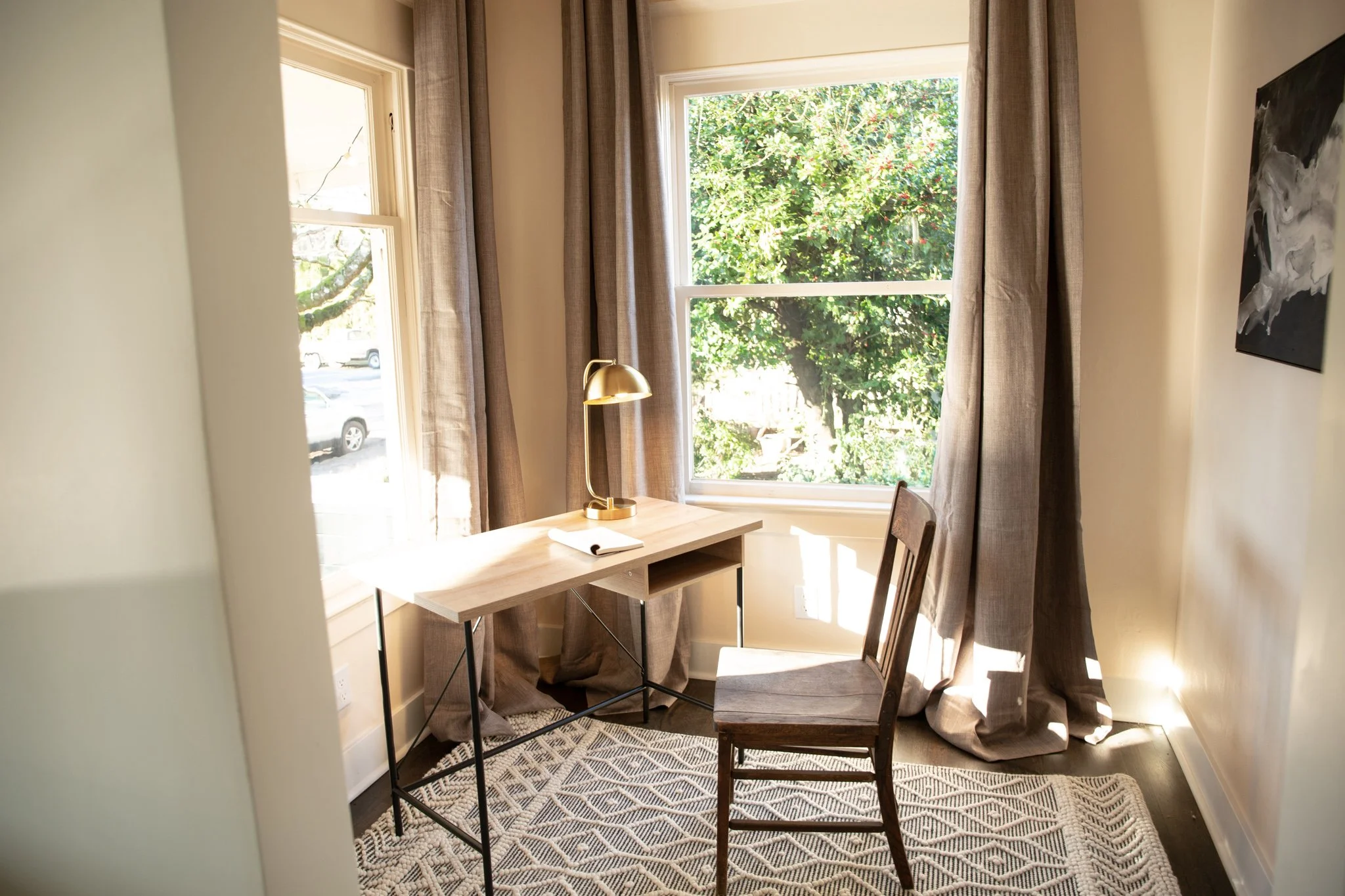 A small home office with a light wood desk, a brown chair, a brass desk lamp, a notebook, and large windows with beige curtains, overlooking green trees outside.