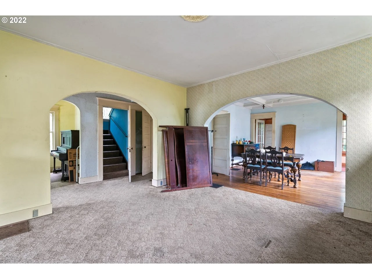 Interior view of a house with a yellow wall, archways, and a mix of carpet and hardwood flooring, featuring a dining area with a dark wood table and chairs.