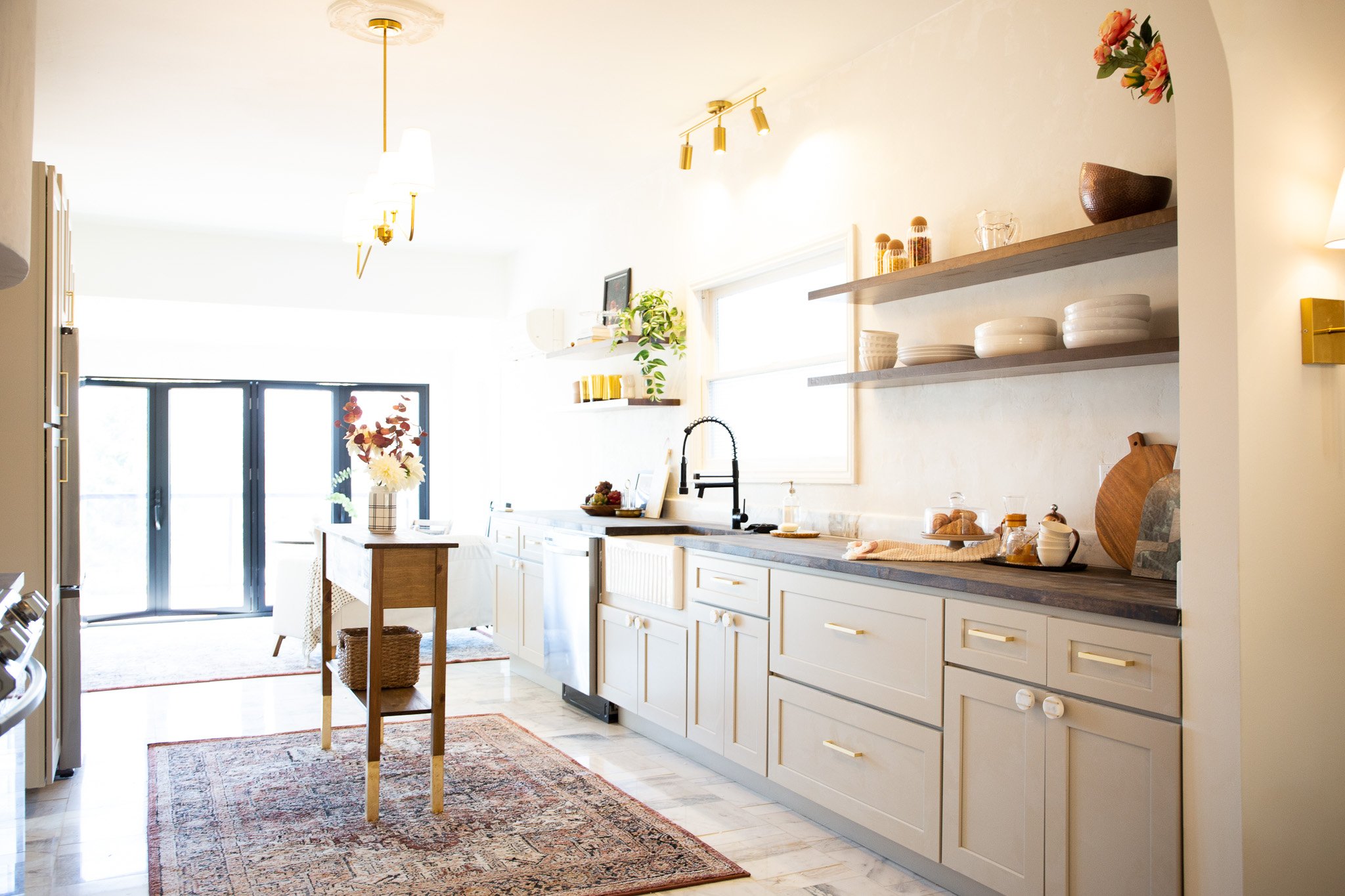 Bright kitchen with white cabinets, open wooden shelves, black sink with a high-arc faucet, a window above the sink, and sliding glass doors at the far end. Decor includes vases with flowers, a bowl of fruit, and various dishes on the shelves and cou