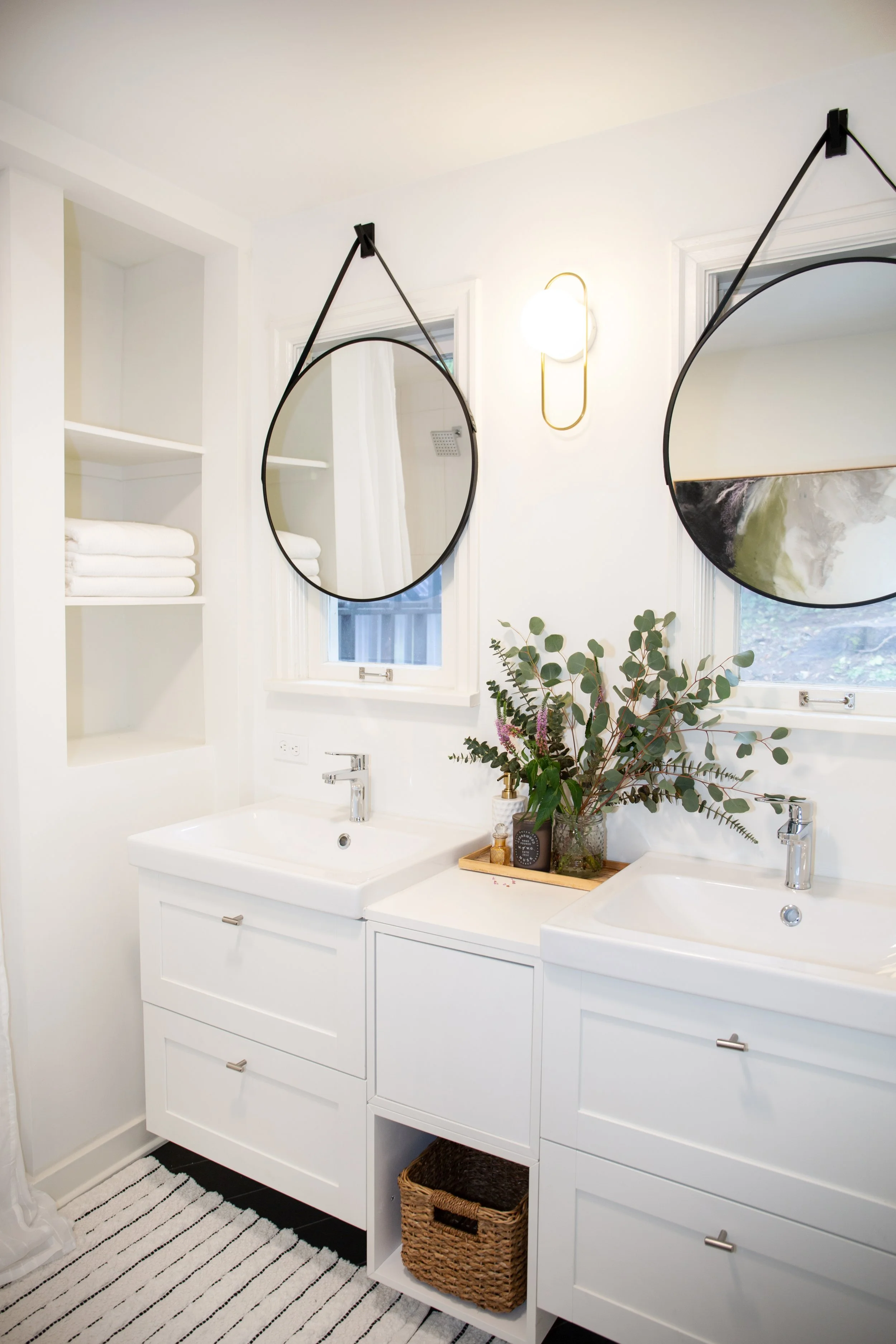 Modern bathroom vanity with double sinks, round mirrors hanging from black straps, open shelving with folded towels, a vase with greenery and flowers, and black and white striped rug.