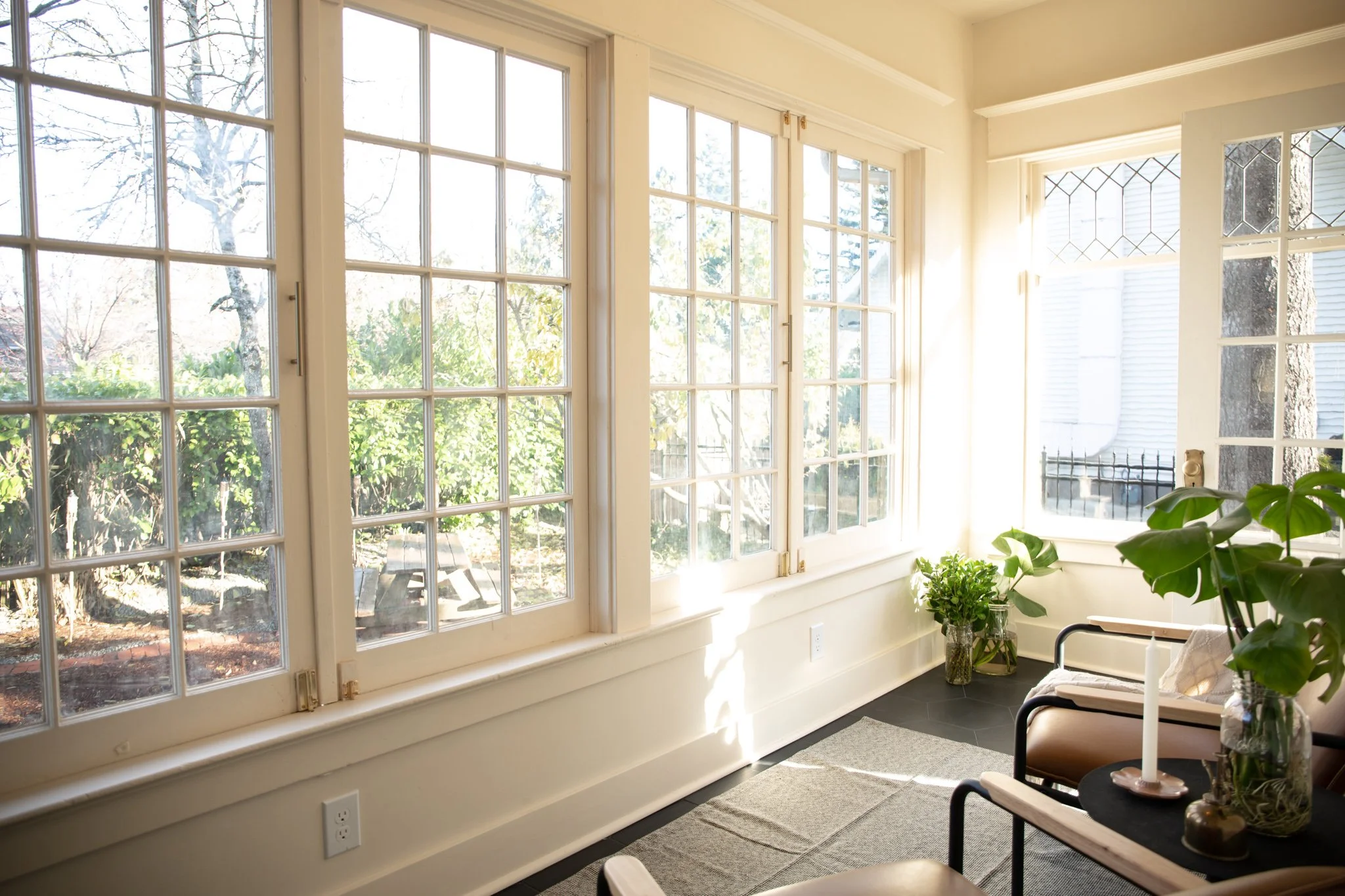 Sunlit room with large windows, potted plants, and a seating area with a candle on a table.