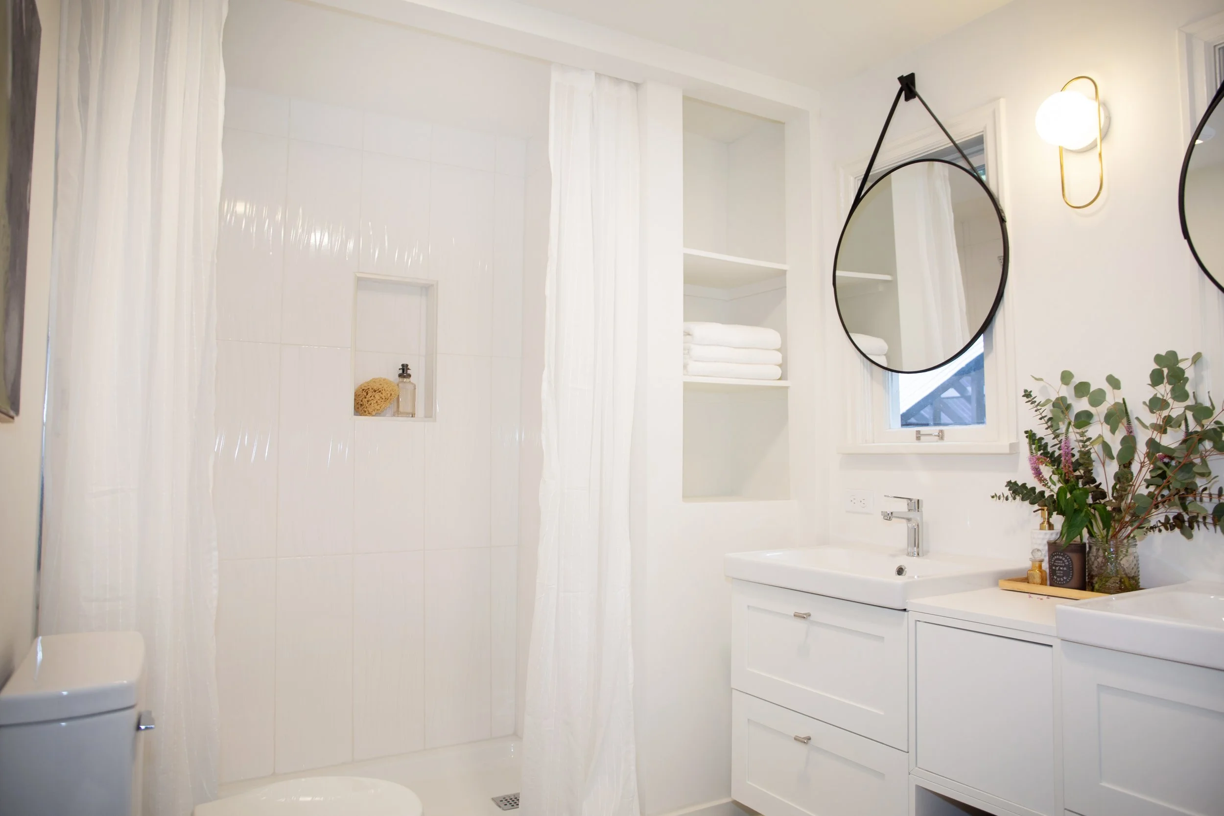 Bright white bathroom with a separate shower area, round mirrors hanging from leather straps, open shelving with folded towels, and a vanity with a sink and a vase of flowers.