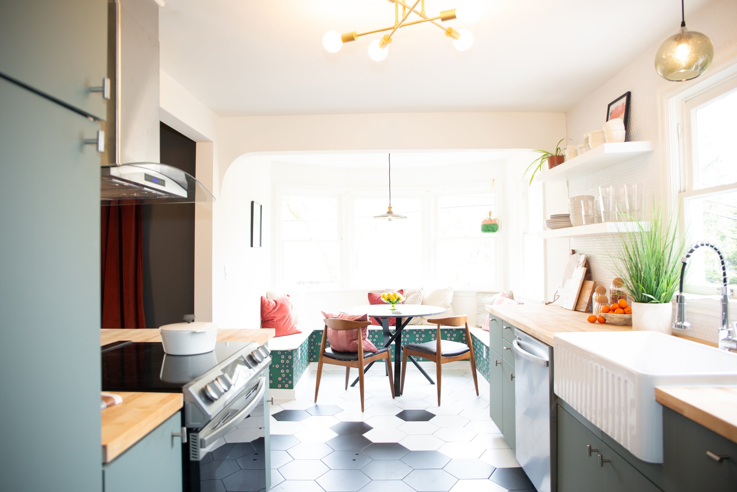 Bright kitchen with a round dining table and chairs near large windows, white walls, pastel cabinets, open shelving with dishes, and a black and white hexagonal tile floor.