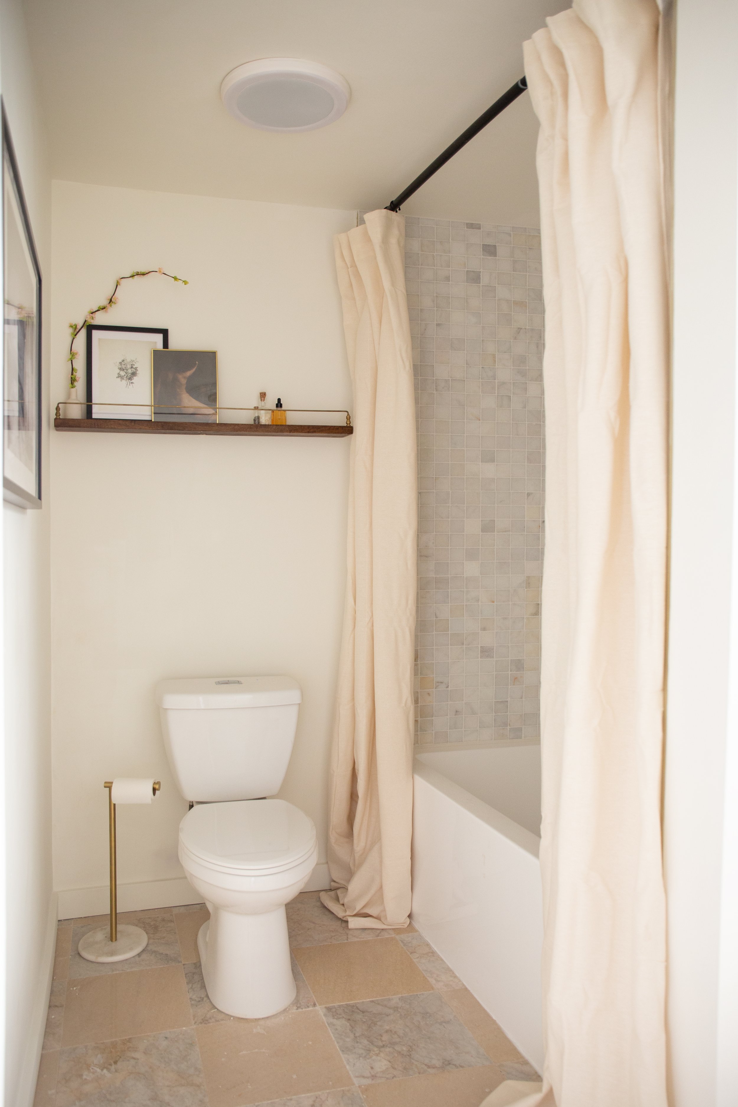 Small bathroom featuring a white toilet, a toilet paper holder, a tiled shower with a beige curtain, and a wooden shelf with framed pictures and decorative items.