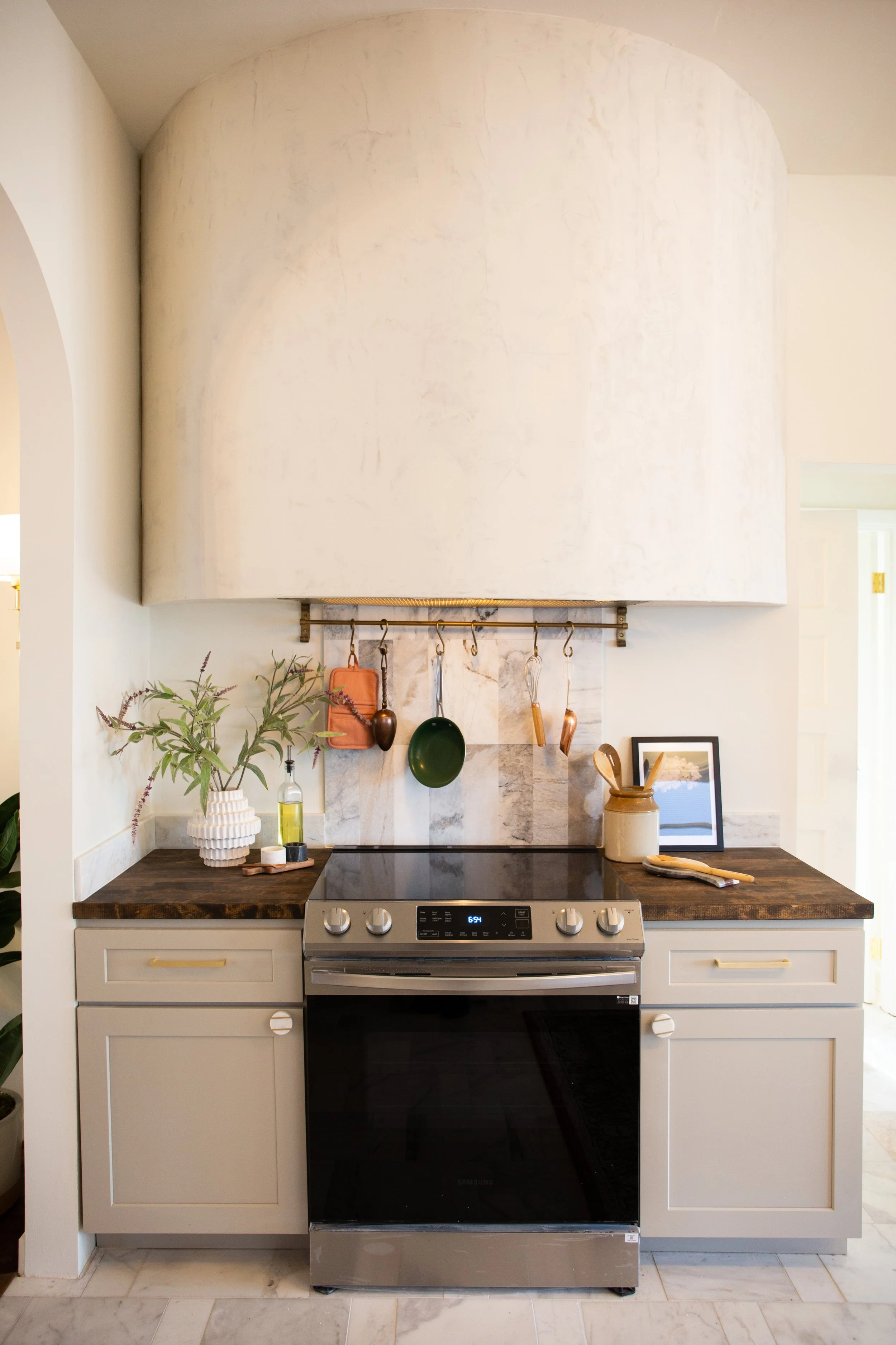 Kitchen with beige cabinets, a dark wooden countertop, a stainless steel oven and stove, decorative items, plants, and hanging utensils.