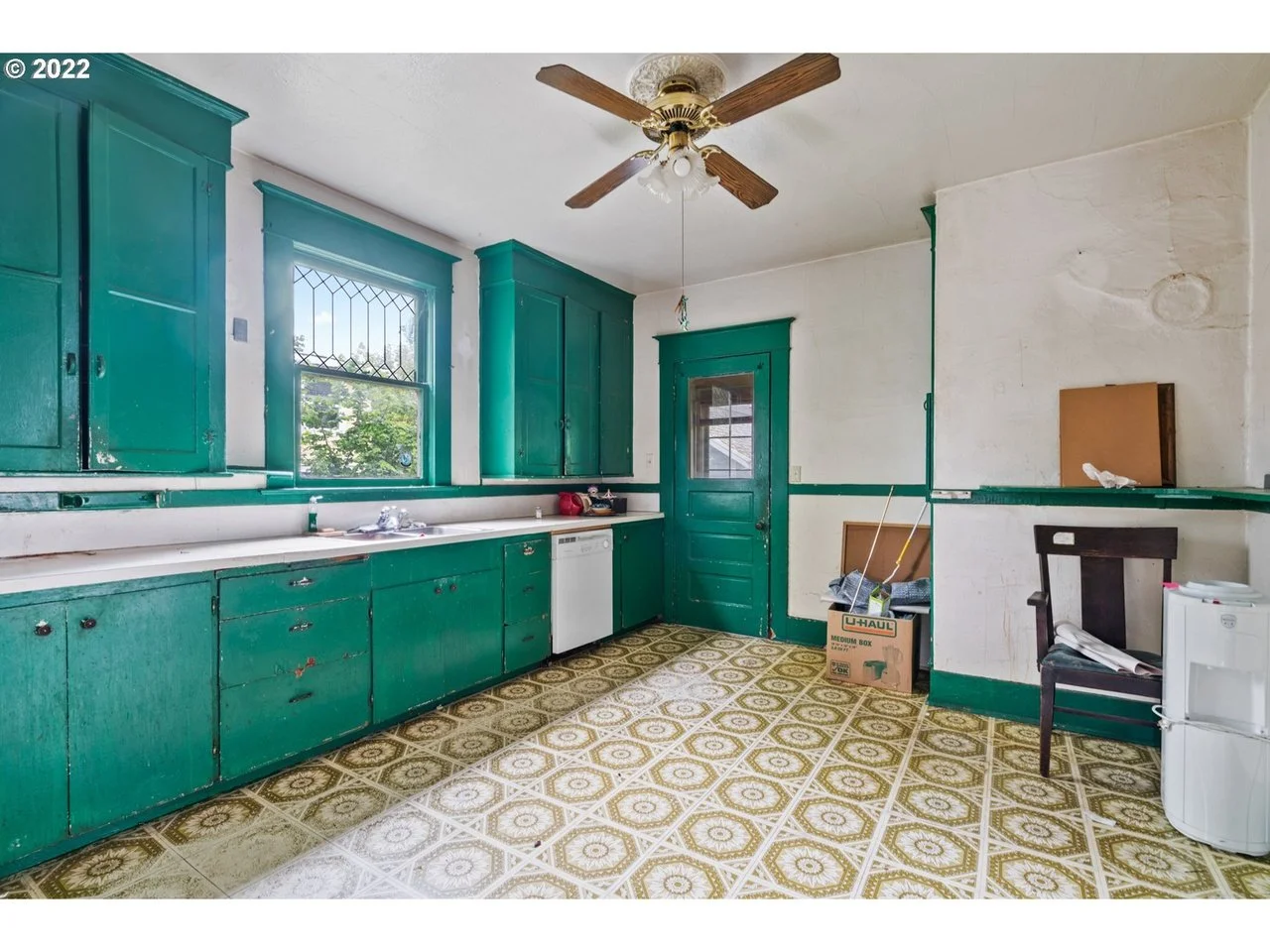 Old kitchen with green cabinets, a window with diamond-shaped panes, a ceiling fan, and patterned yellow and brown linoleum flooring. Various items including a cardboard box, a chair, and a water dispenser are visible.