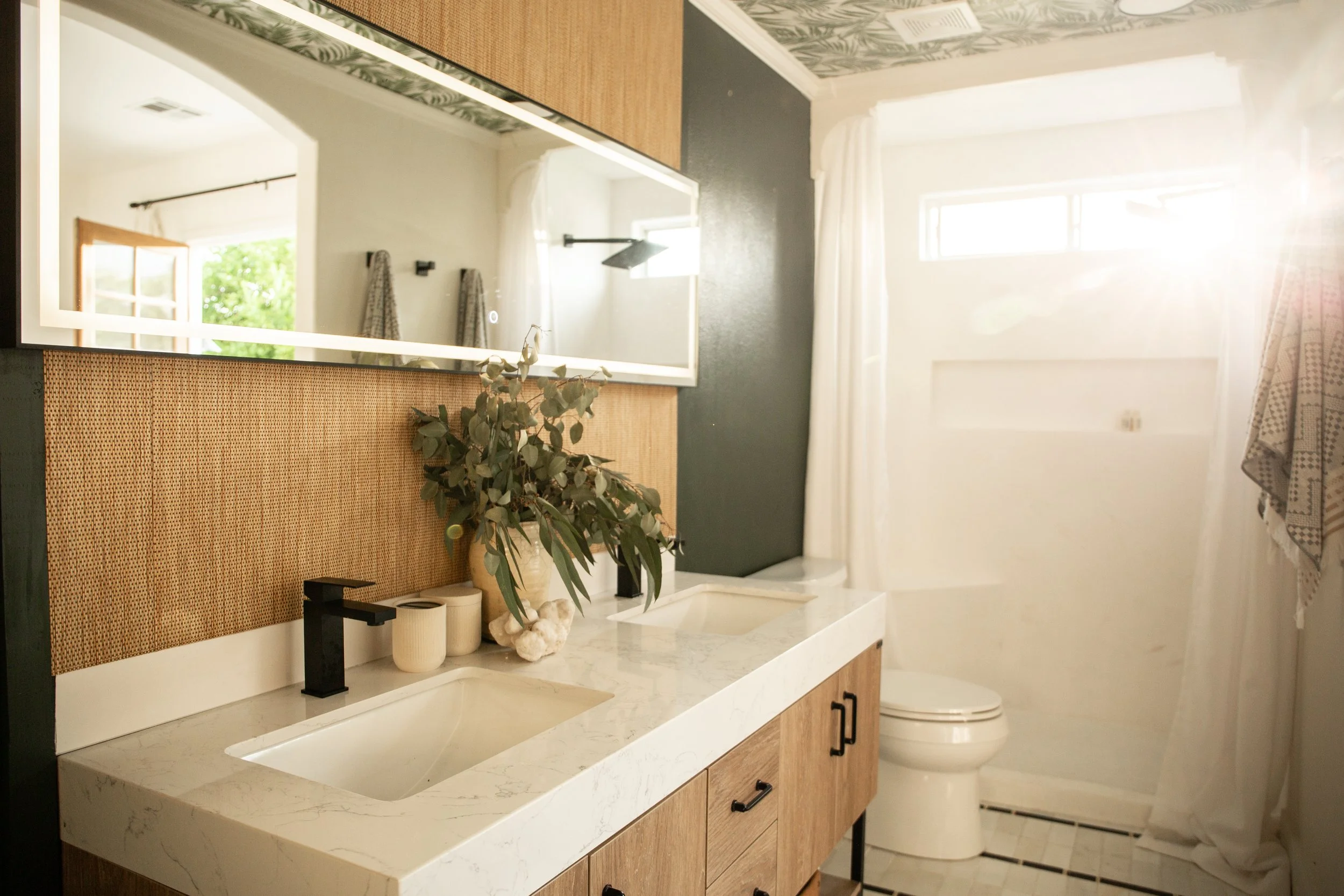 A bathroom with a double sink vanity, a large mirror, and a toilet. The vanity has a white marble countertop with two integrated sinks, black faucets, and wooden cabinets. There are decorative items including a vase with greenery and candles. The bac