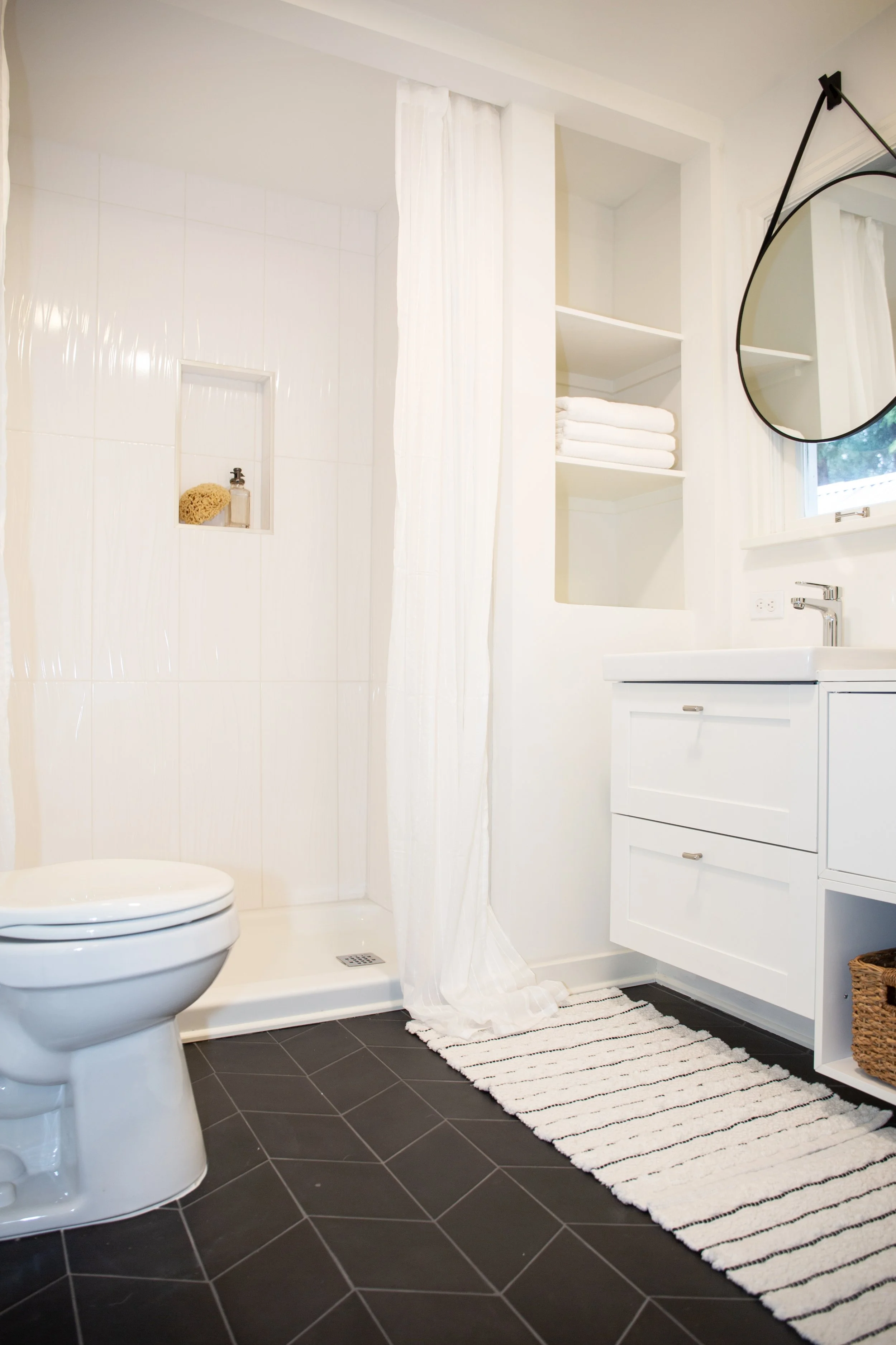A clean bathroom featuring a white toilet, an uncluttered shower with a built-in niche holding a sponge and soap, a white vanity with drawers, a round mirror hanging from a black strap, a striped rug on dark tiled flooring, and open shelves with neat