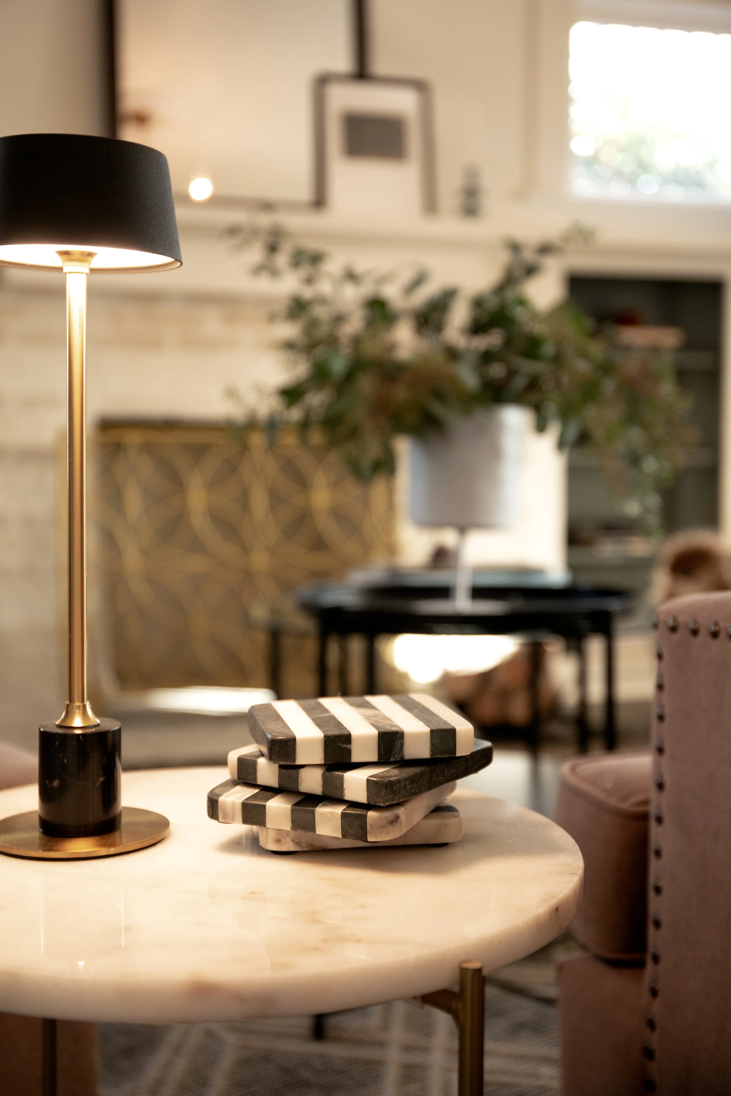 A stack of black and white striped coasters on a round marble table, with a modern table lamp to the left, in a cozy living room with a potted plant and decorative artwork in the background.