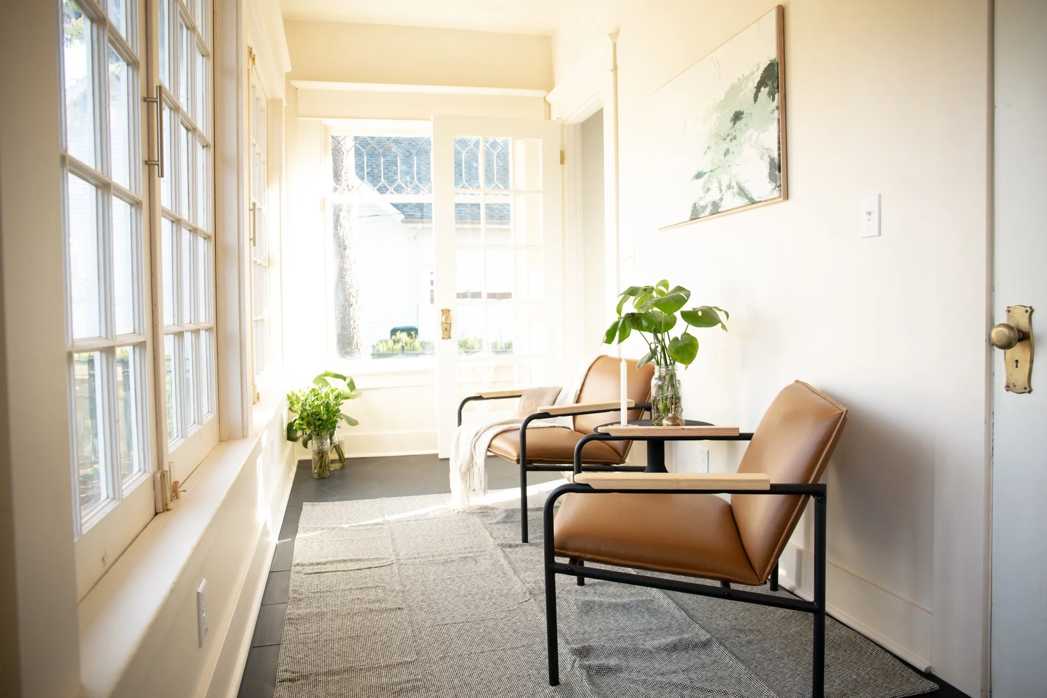 A cozy sitting area with two tan leather chairs, small wooden tables, and green potted plants. Sunlight streams through large windows with white frames, illuminating the space.