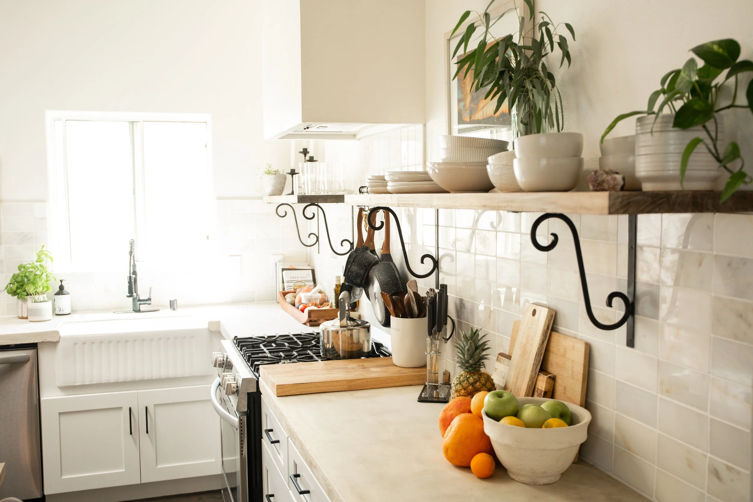 Bright kitchen with a window, white cabinetry, a gas stove, and open wooden shelves holding bowls, plates, and potted plants. Countertop contains a cutting board, a bowl of various fruits, and utensils.