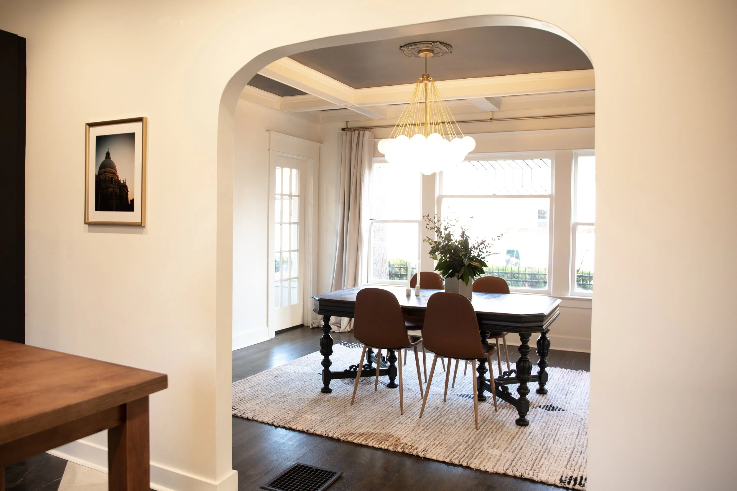 Dining room with a wooden table, four brown chairs, a floral centerpiece, large windows, white curtains, a modern chandelier, and a beige rug.