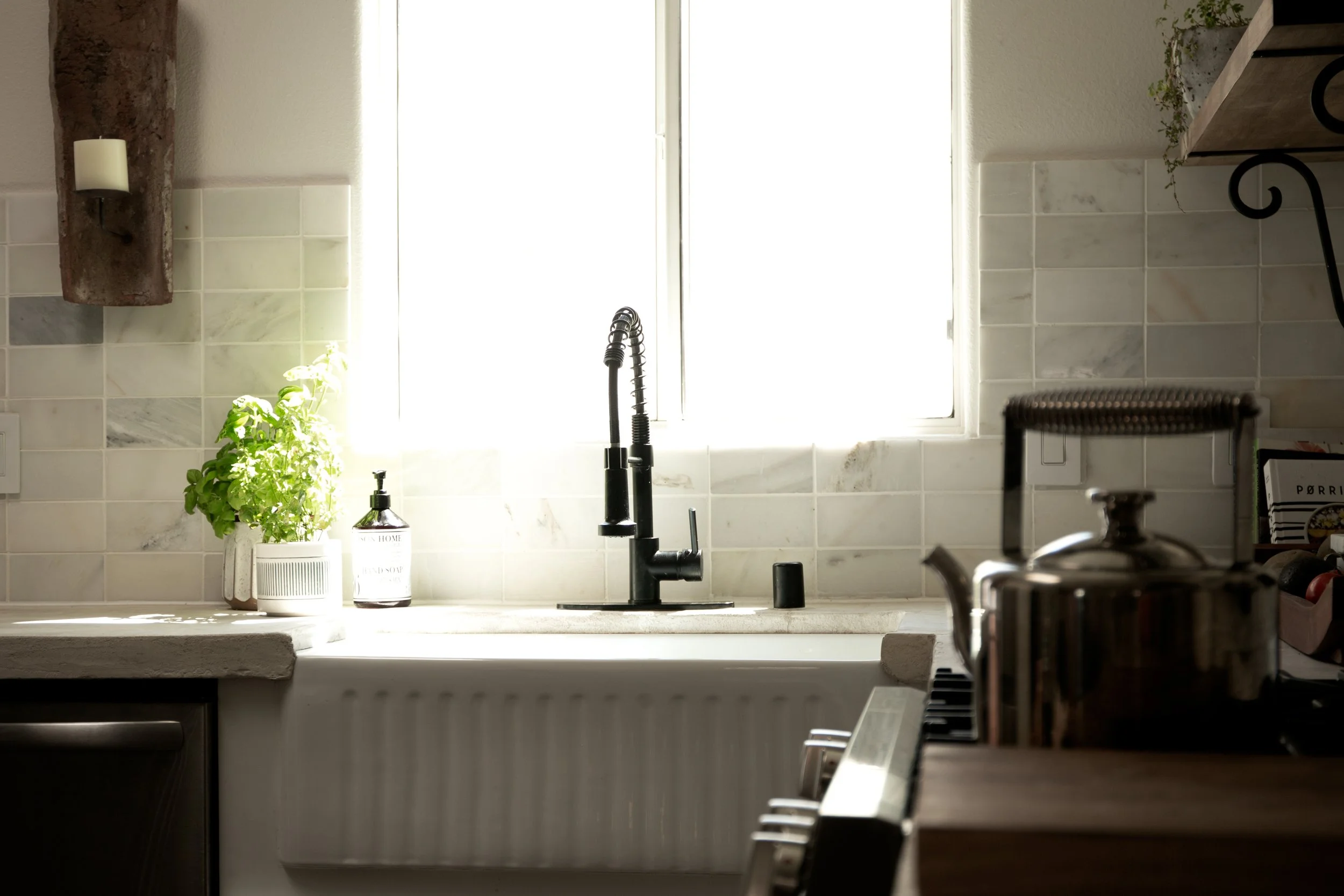 Kitchen with window, black sink faucet, potted green plant, soap dispenser, stove with a kettle, and tiled backsplash.