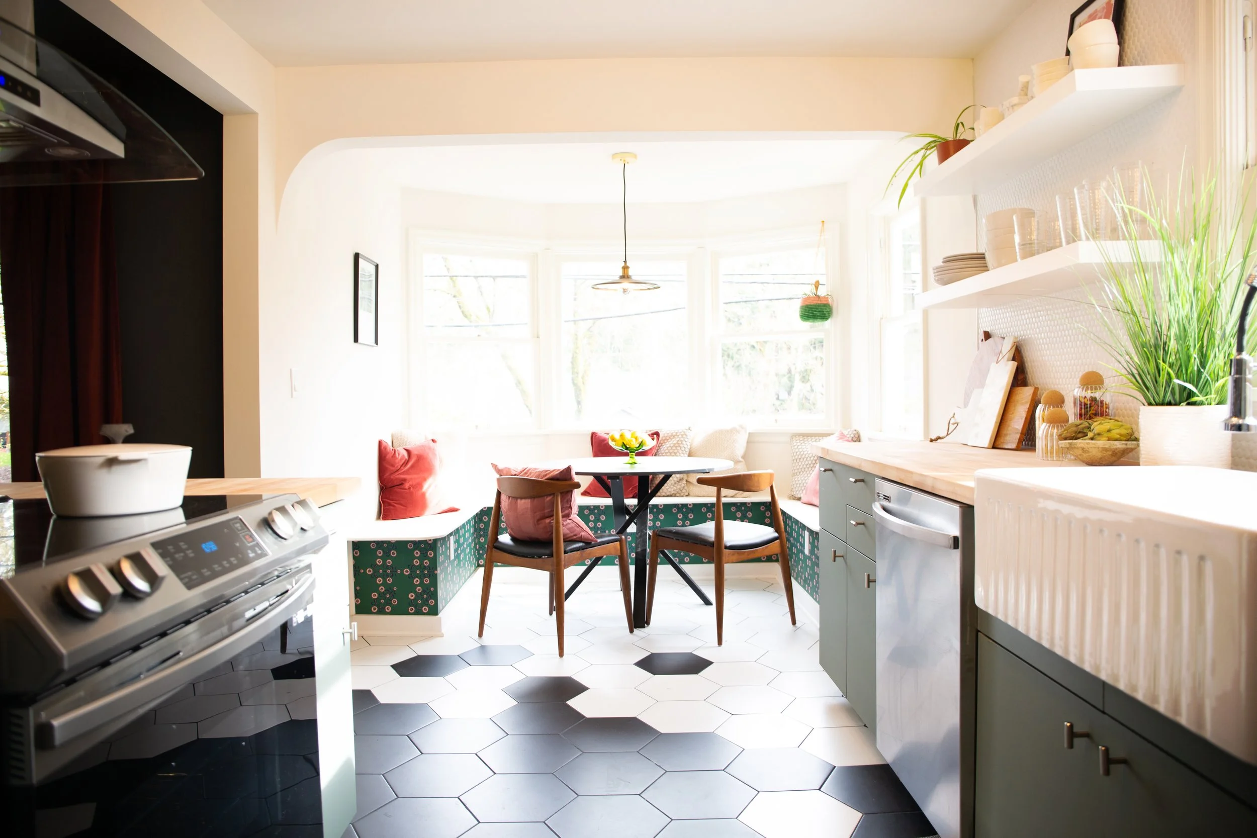 Bright kitchen with a dining nook near large bay windows, featuring a round table with four chairs and built-in bench seating with red and white pillows. Cabinets and countertops line one wall, with open shelves holding dishes, plants, and decorative