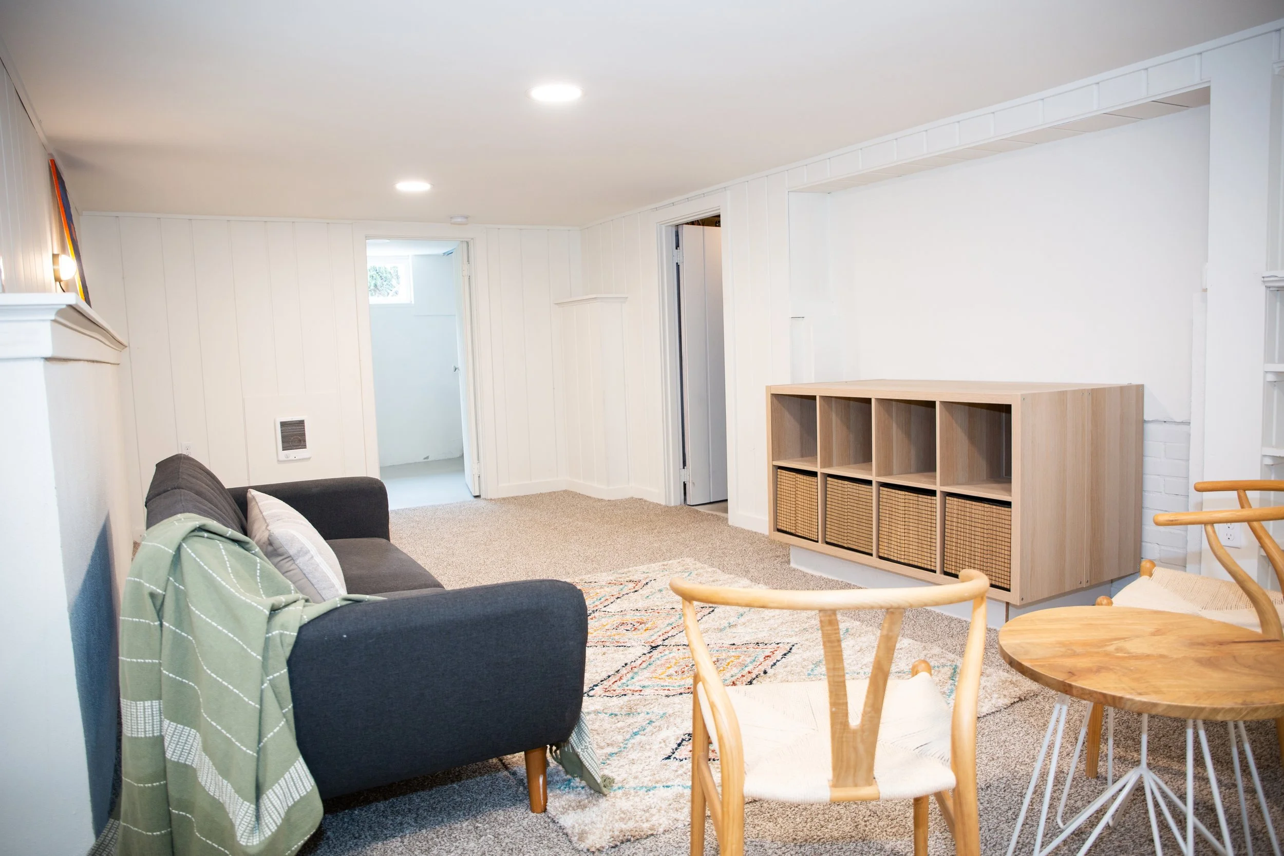 A cozy living room with a gray sofa, a wooden bookshelf with baskets, a round wooden table, and a small seating area in a finished basement with white walls and beige carpet.