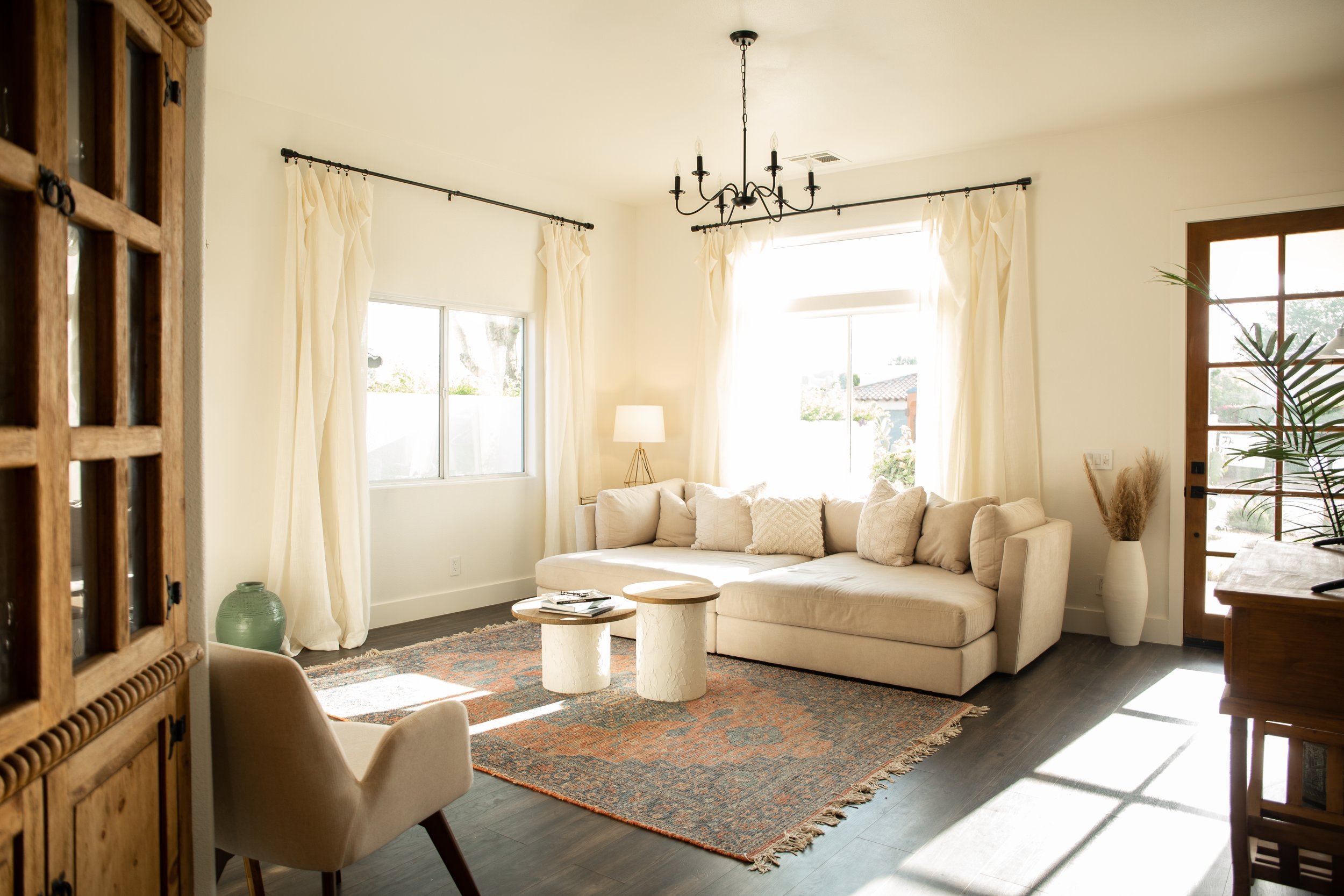 Bright living room with cream-colored sectional sofa, patterned area rug, two small round coffee tables, a table lamp, potted plants, and large windows with cream curtains.