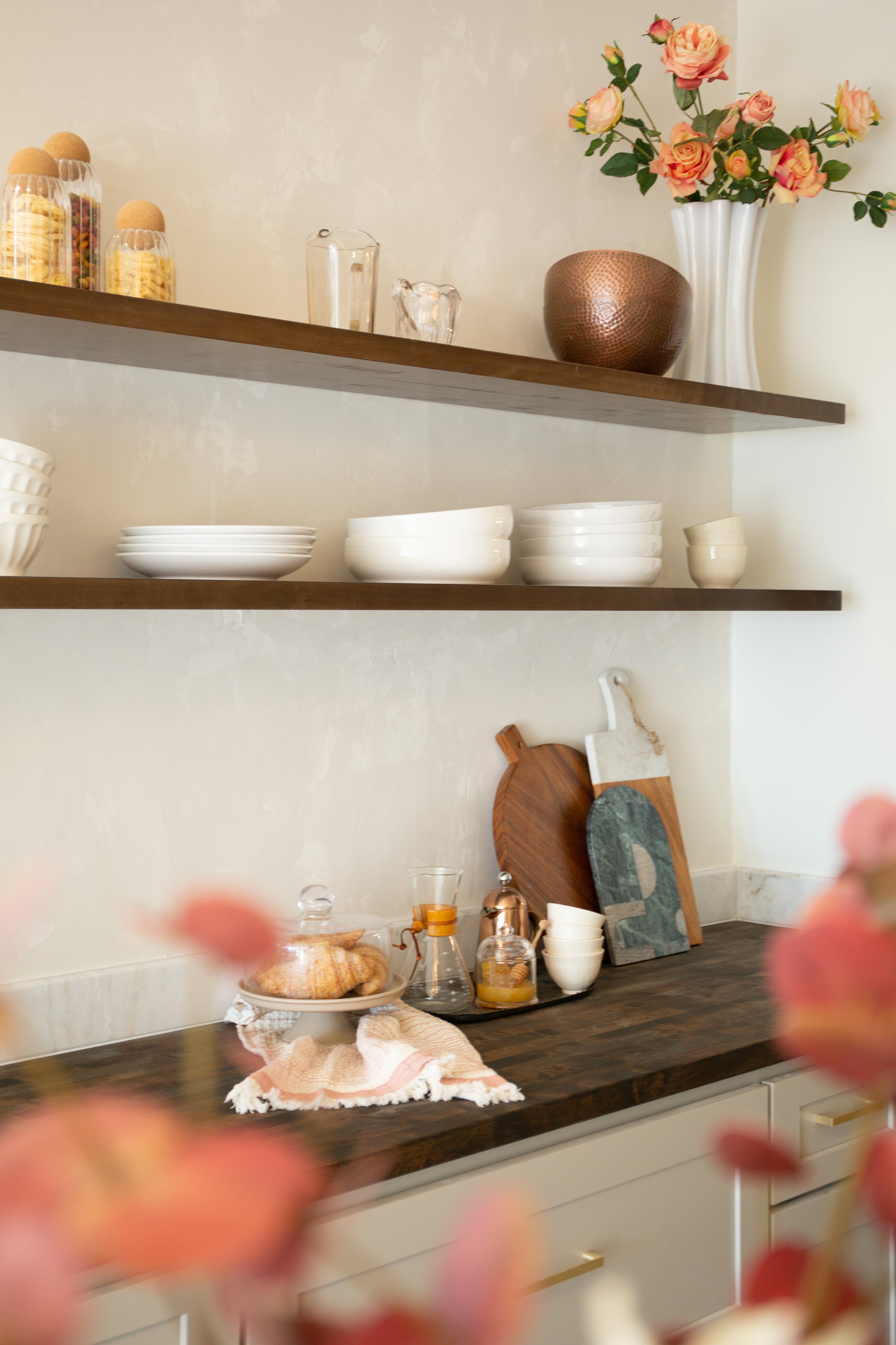 Kitchen shelf with glass jars of snacks, a white vase with pink roses, and decorative items. On the counter, there are cutting boards, glass bottles, a covered pastry, and small bowls, with pink flowers in the foreground.