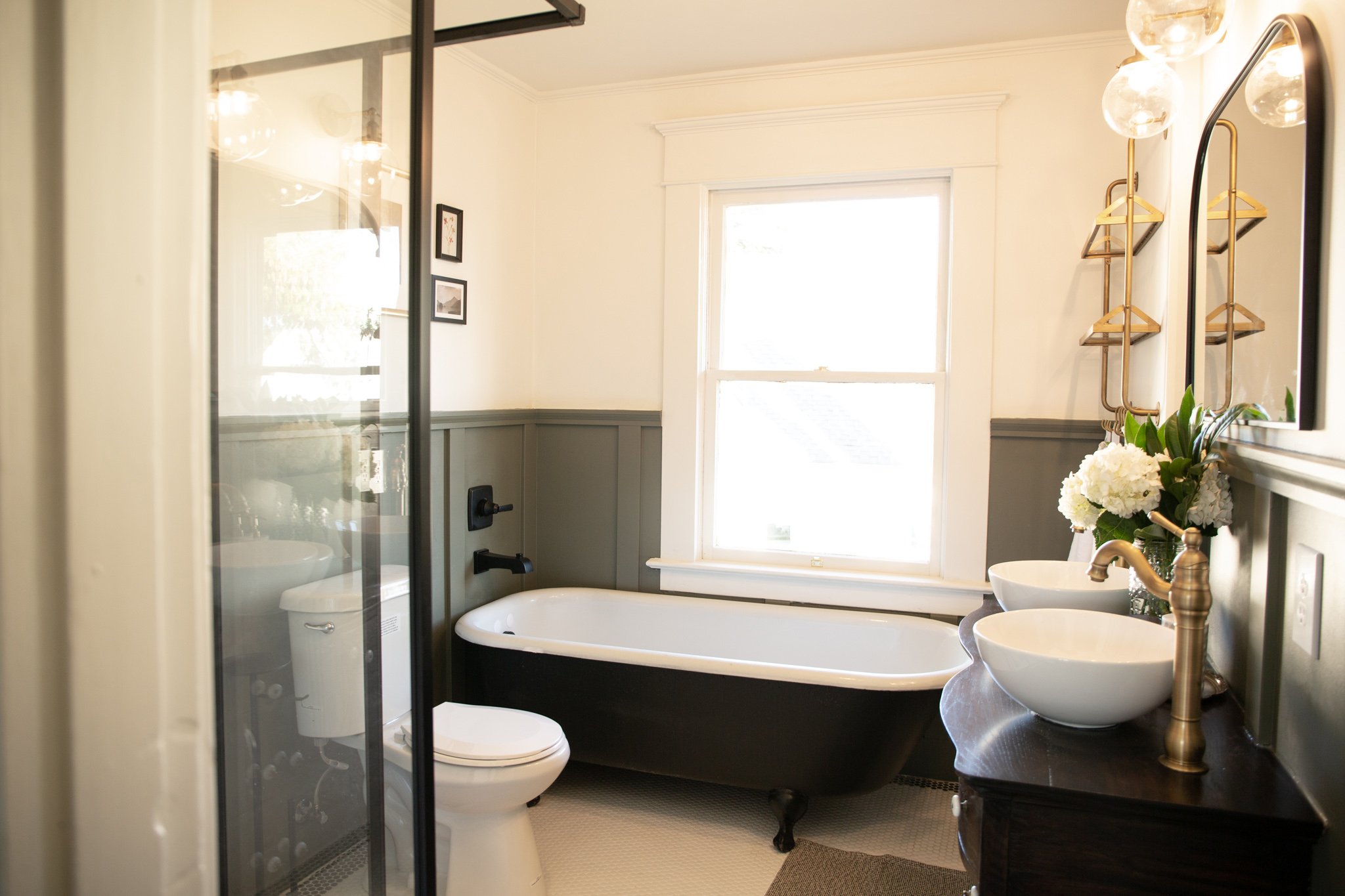 A bathroom featuring a black clawfoot tub, a double sink vanity with white vessel sinks, a mirror, a window, and decorative flowers.