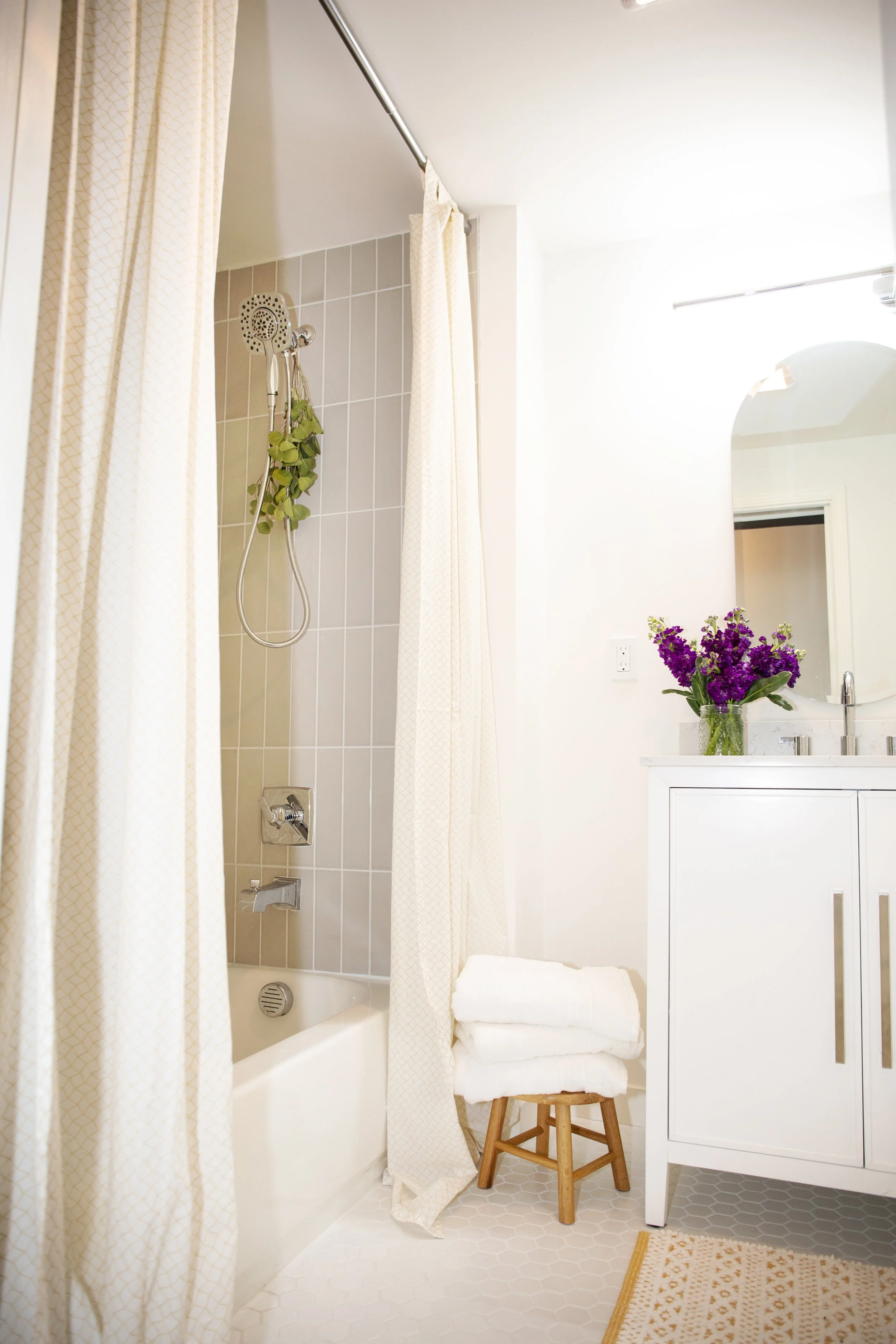 A bathroom with a shower area, beige tile walls, a white bathtub, off-white shower curtain, a white vanity with a silver faucet, a mirror, a vase with purple flowers, a wooden stool with folded white towels, and a patterned rug on the white hexagonal