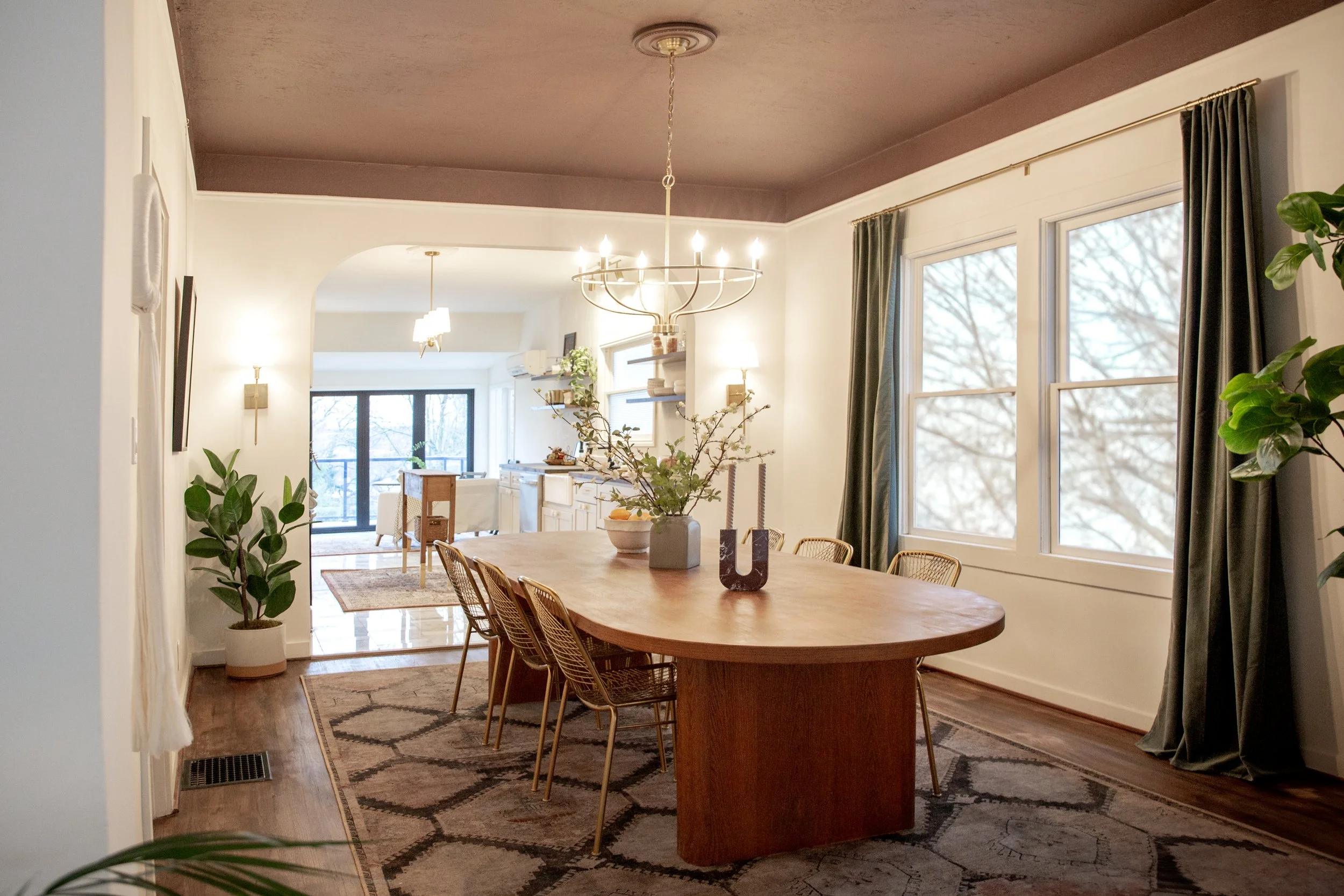 A cozy dining room with a large wooden table, surrounded by gold-colored wire chairs. A chandelier hangs above the table, decorated with a floral arrangement. Large windows with dark curtains admit natural light, revealing a snowy outdoor scene. The 