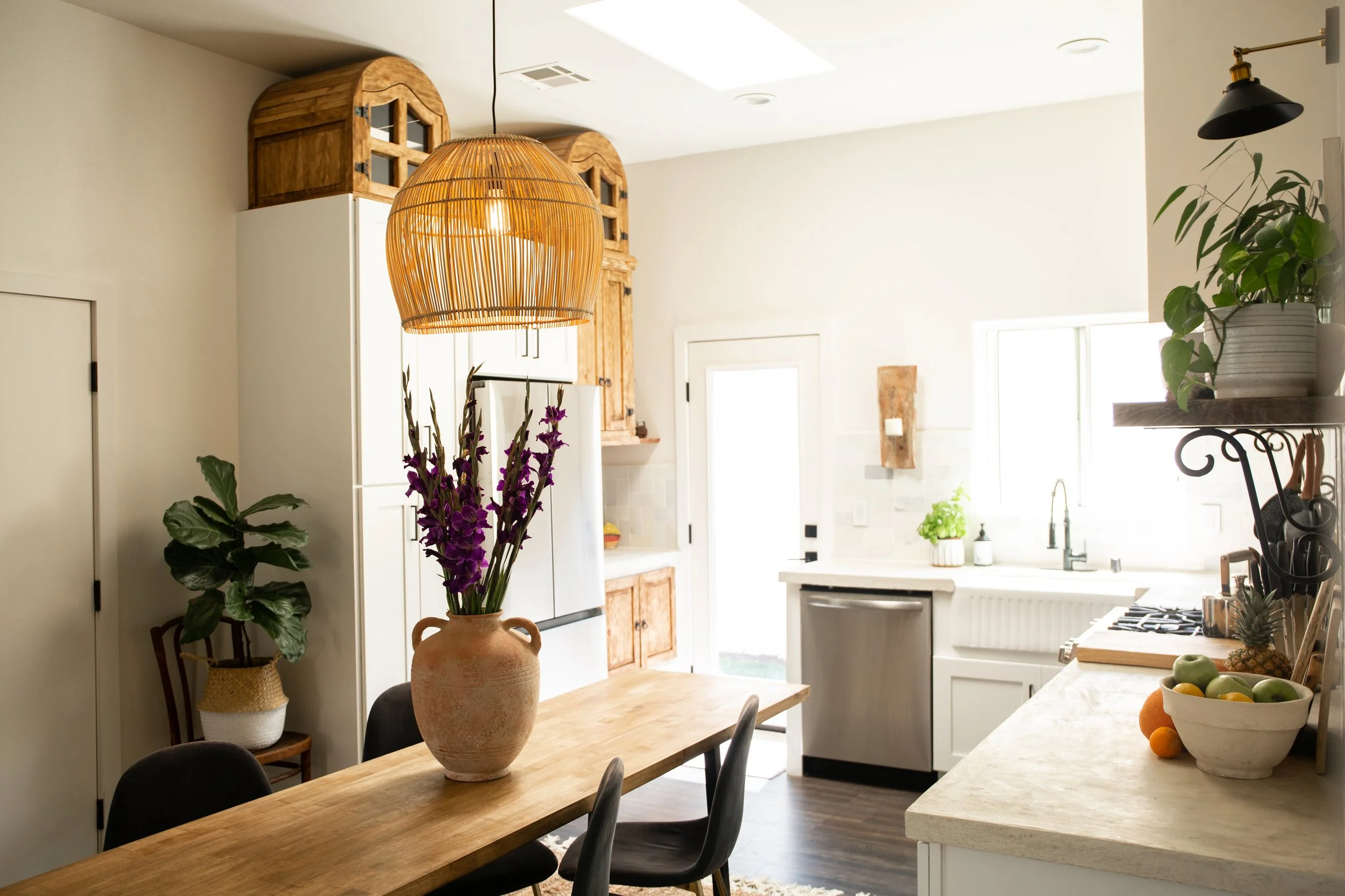 Bright kitchen with wooden dining table, potted plants, a hanging wicker light, and white cabinets