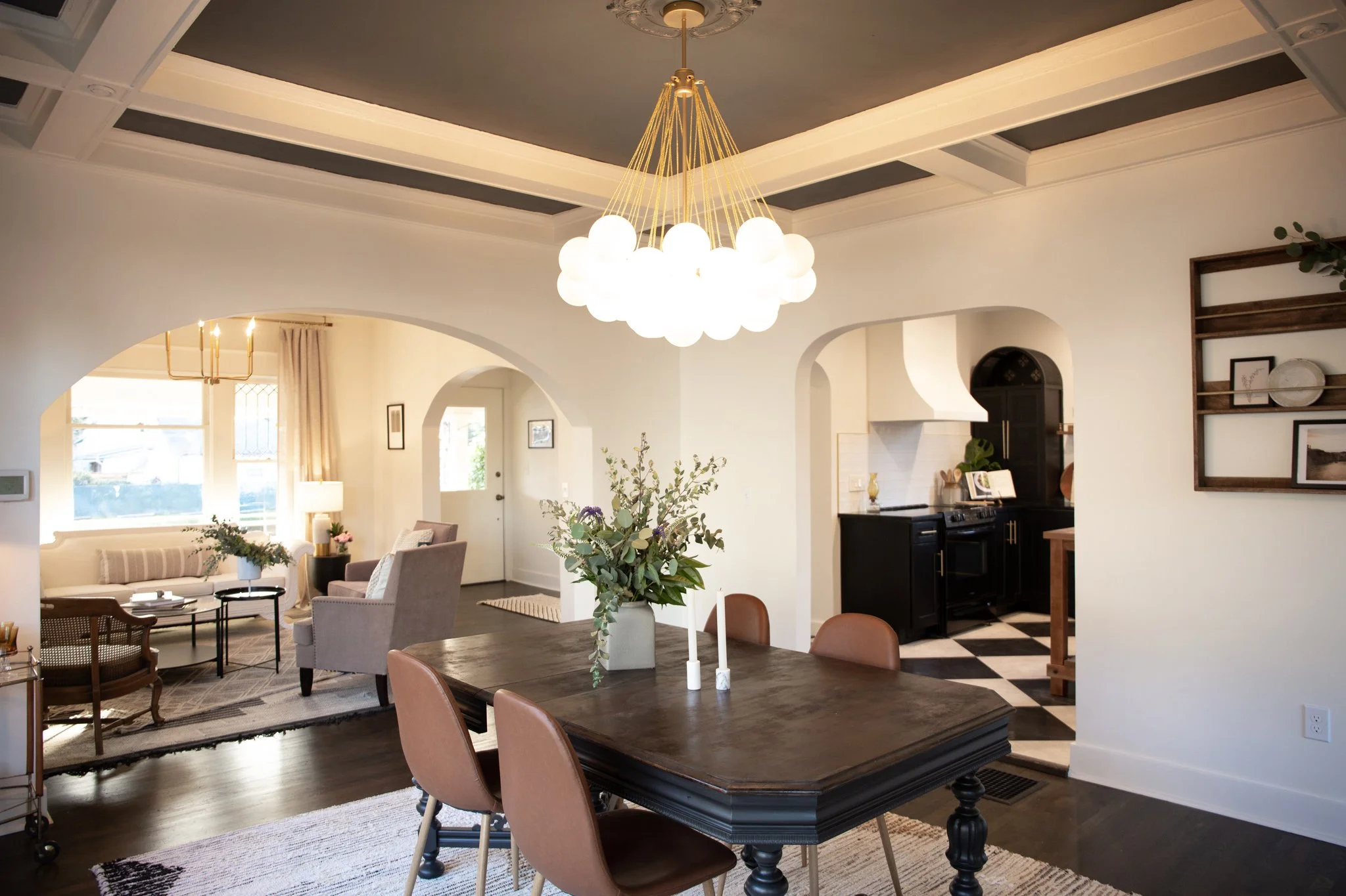 Open-concept living and dining room with a dark wood dining table, pink chairs, chandelier, and a cozy sitting area with armchairs and sofa, leading to a kitchen with black cabinets and checkered floor, decorated in a neutral color palette.