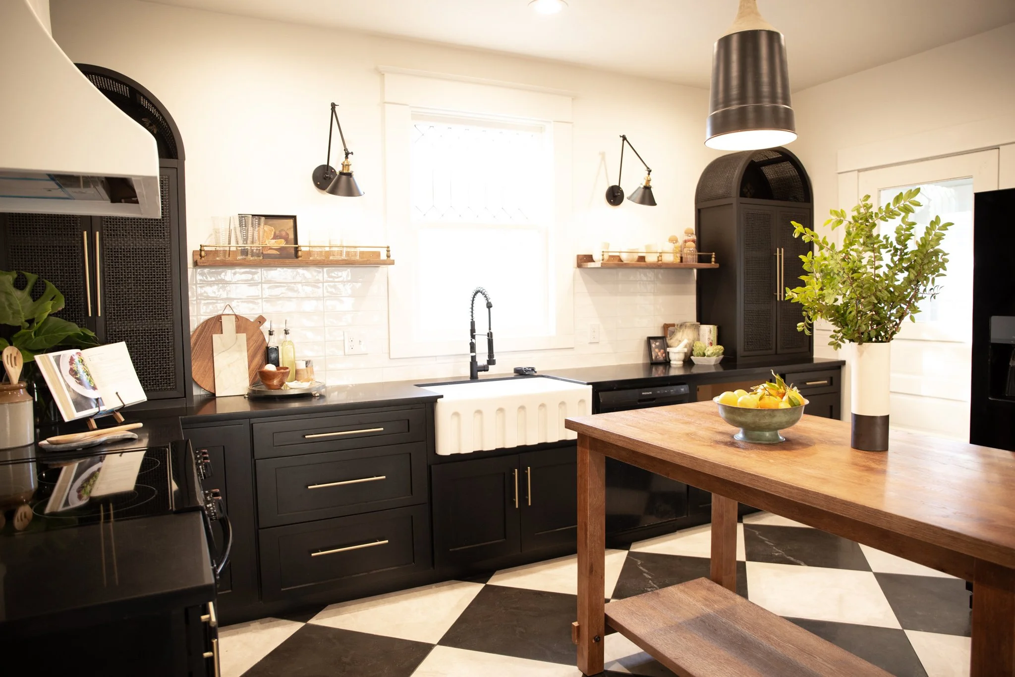 Modern kitchen with black cabinets, white subway tile backsplash, a white farmhouse sink, black fixtures, open wooden shelves, a wooden table with a bowl of lemons, a potted plant, and black and white checkered flooring.
