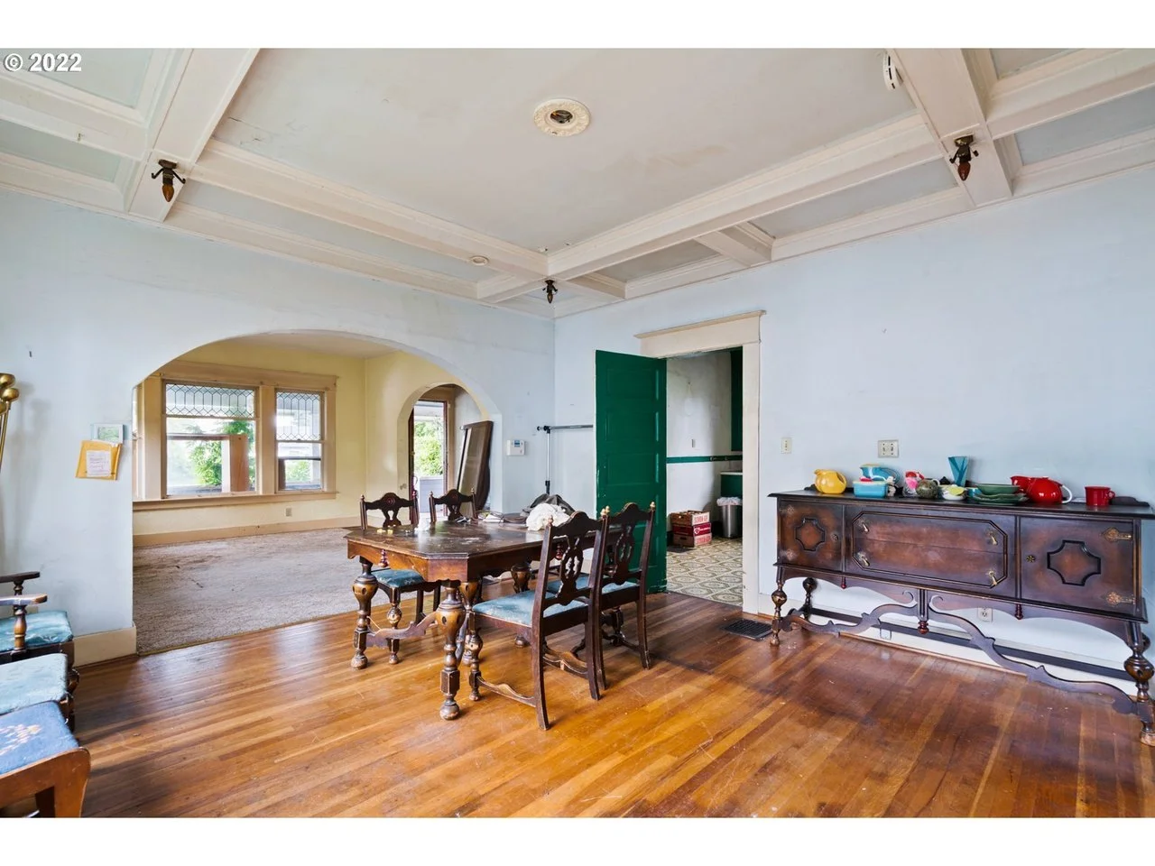 A dining room with hardwood floors, a wooden dining table with chairs, a sideboard with dishes, and an open doorway leading to another room with wallpapered floor.