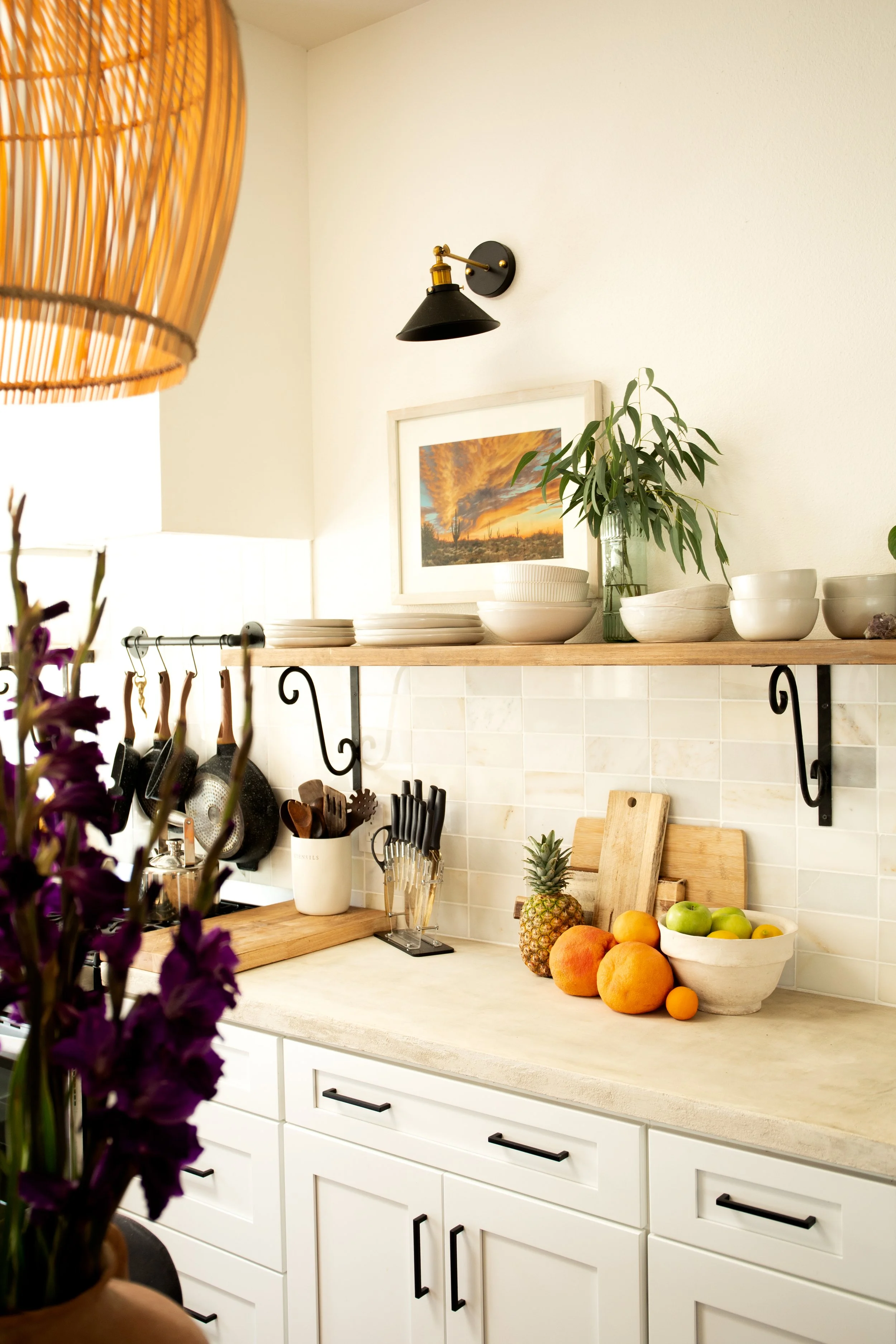 Kitchen with white cabinets, a beige countertop, and a backsplash made of small cream tiles. A wooden shelf holds white dishes, bowls, and a vase with green leaves. Countertop has a pineapple, oranges, apples, and a white bowl of fruit, along with cu