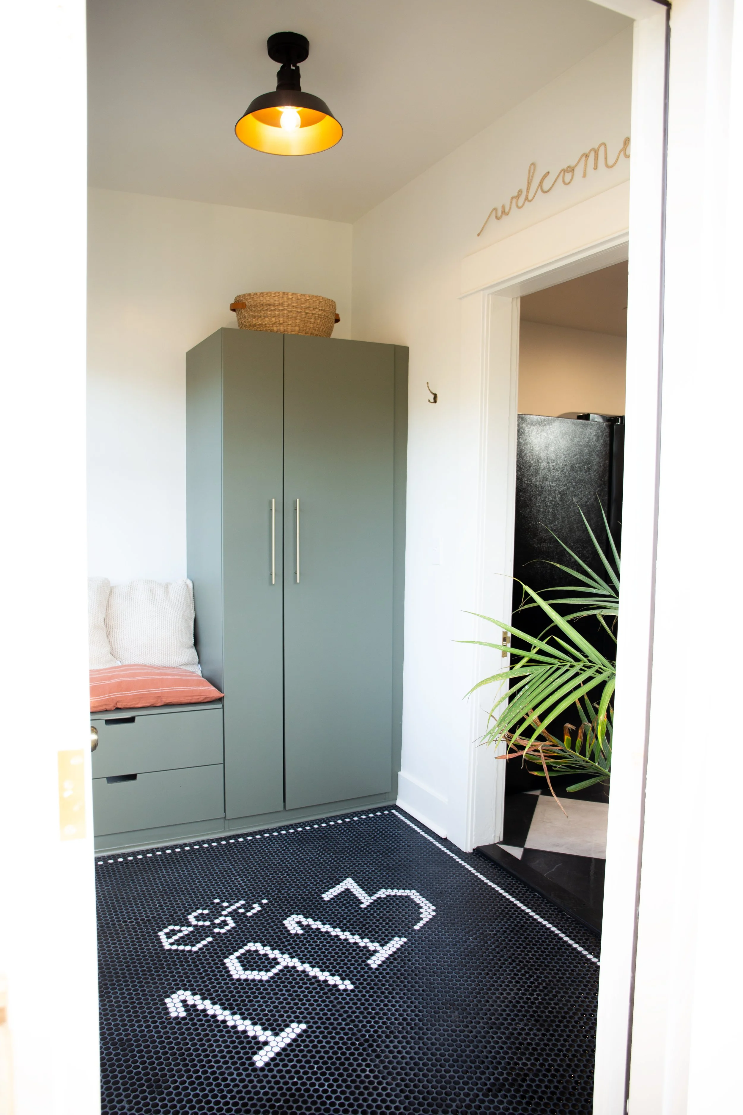 Entryway with a black hexagonal tile rug with white pattern, a green storage bench with pillows, a green cabinet, and a wall sign that says "welcome."