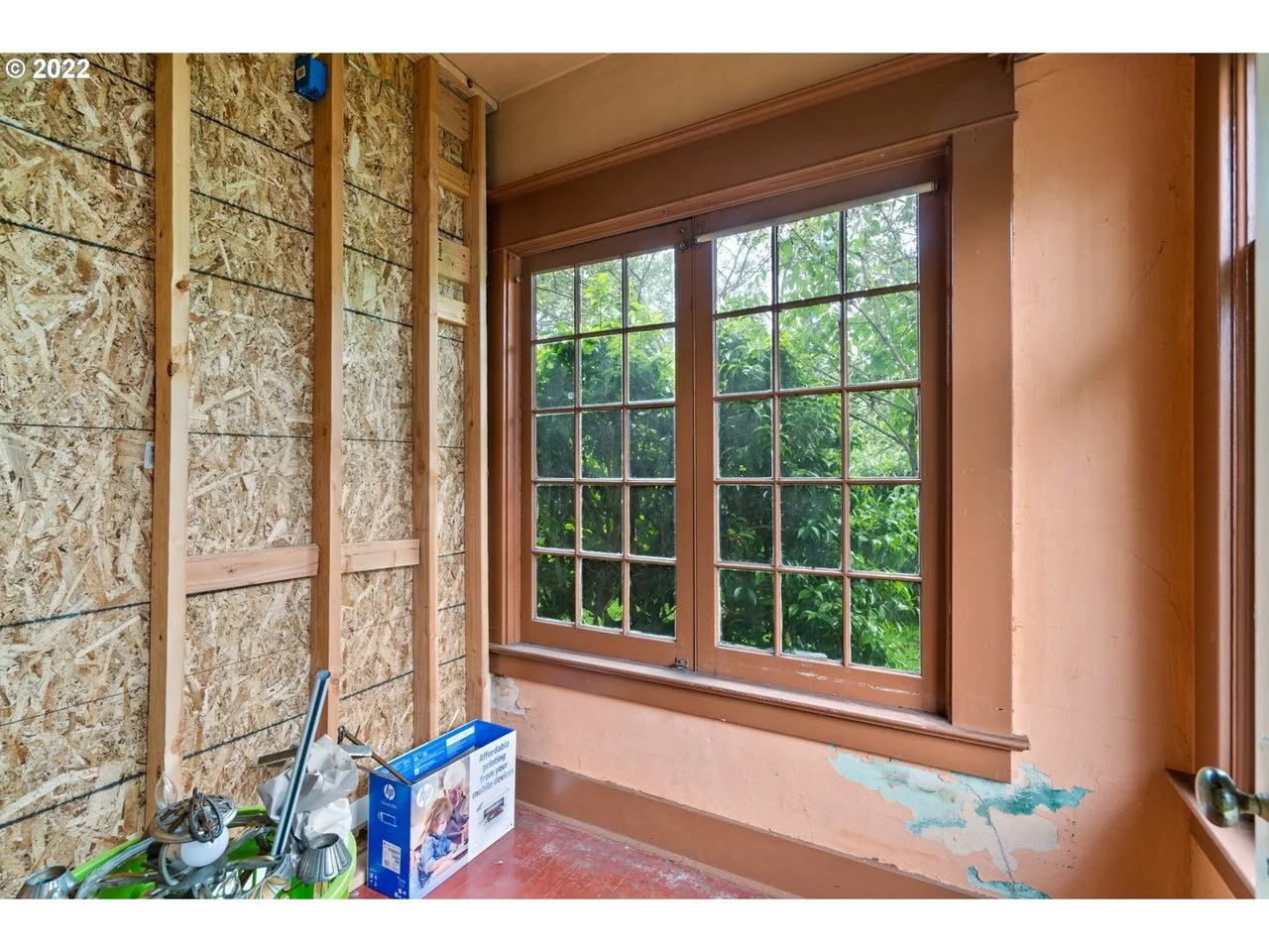 Room under renovation with unfinished wall on the left, a large window with multiple panes on the right showing green trees outside, and peeling pinkish-orange paint on the wall below the window. Construction tools and a box are on the floor.
