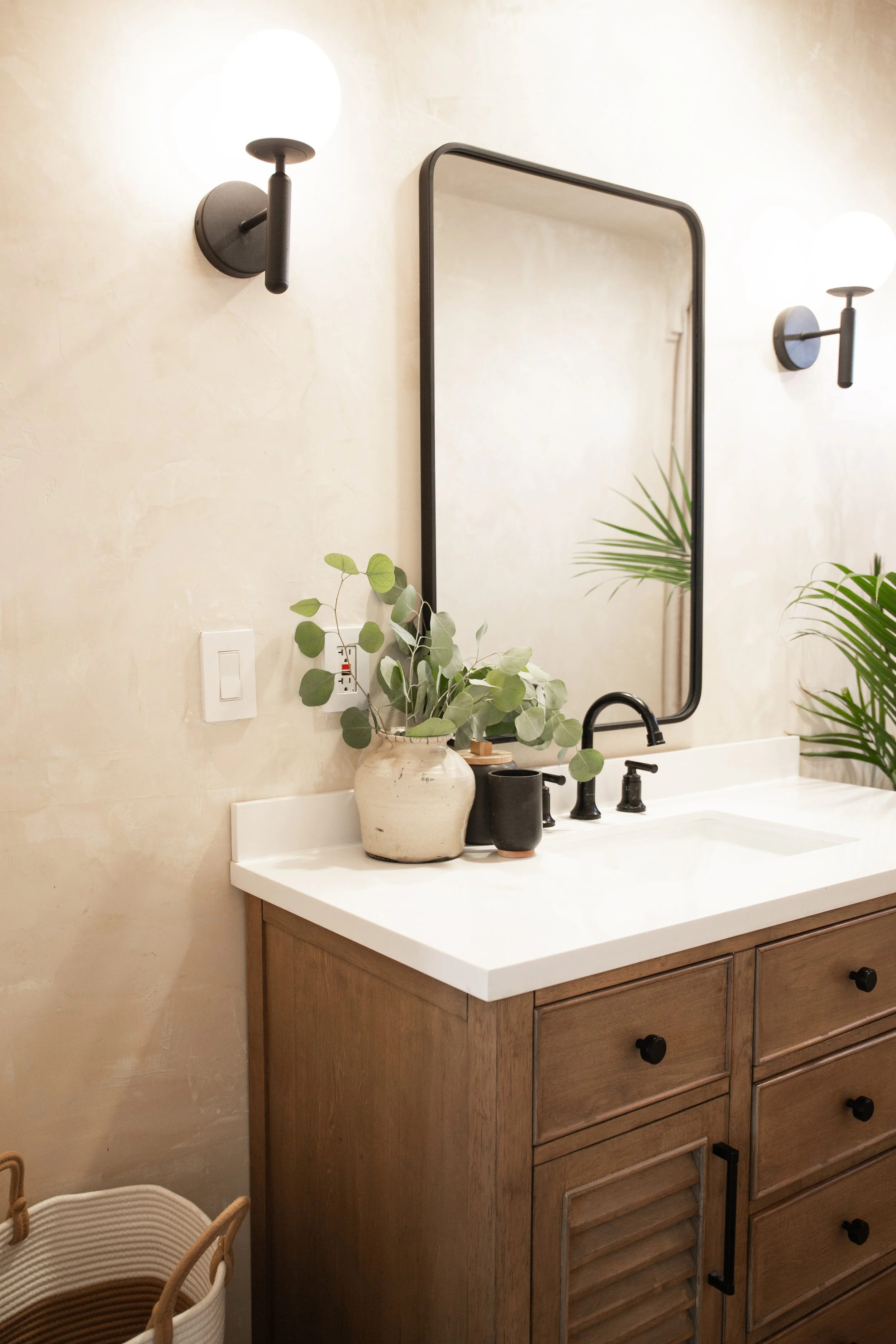 Bathroom vanity with a white countertop, black faucet, mirror, decorative plants in pots, and black wall-mounted light fixtures.