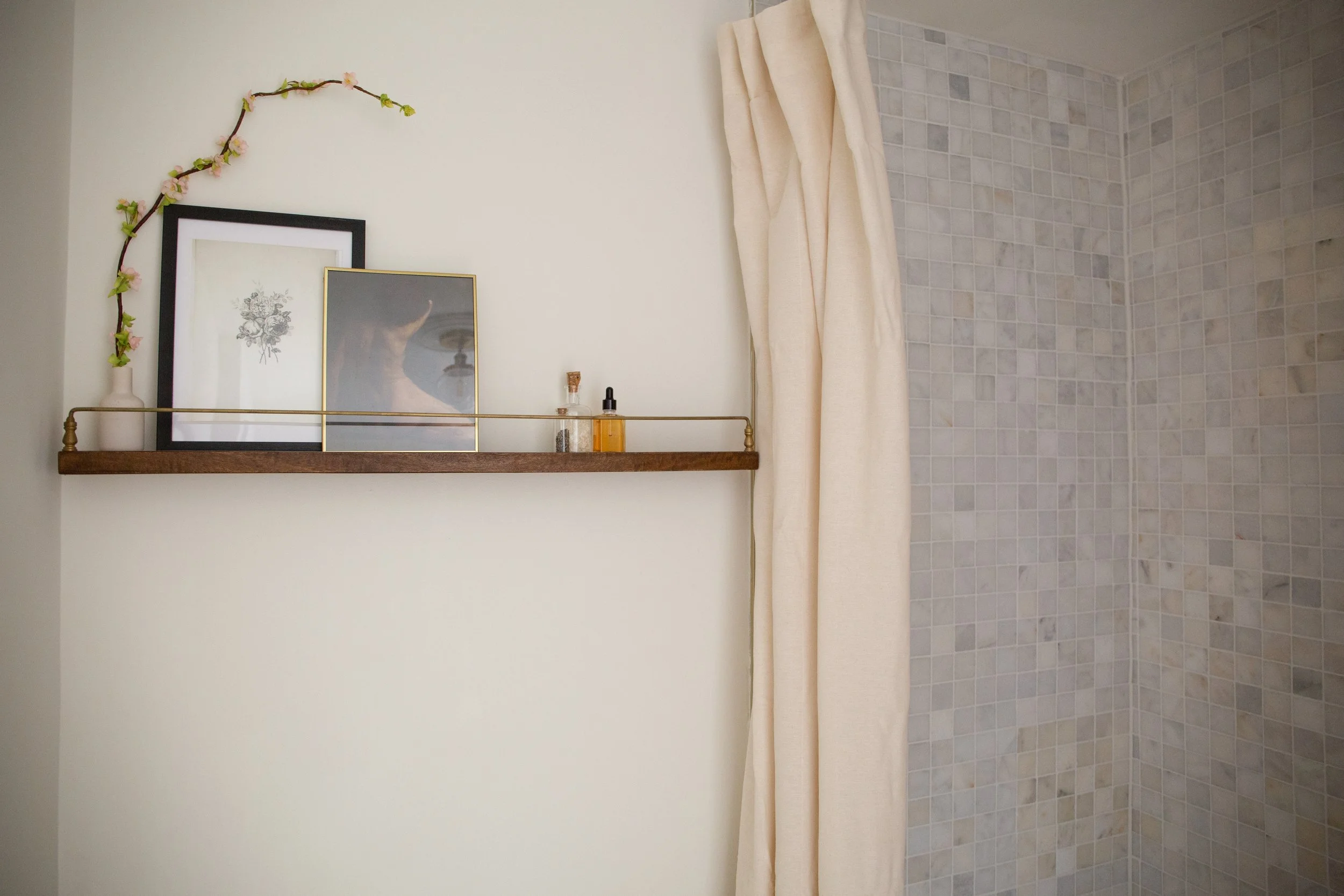 Decorative bathroom shelf with framed artwork, a vase with a branch of pink flowers, and small bottles, next to a tiled shower area with a cream curtain.
