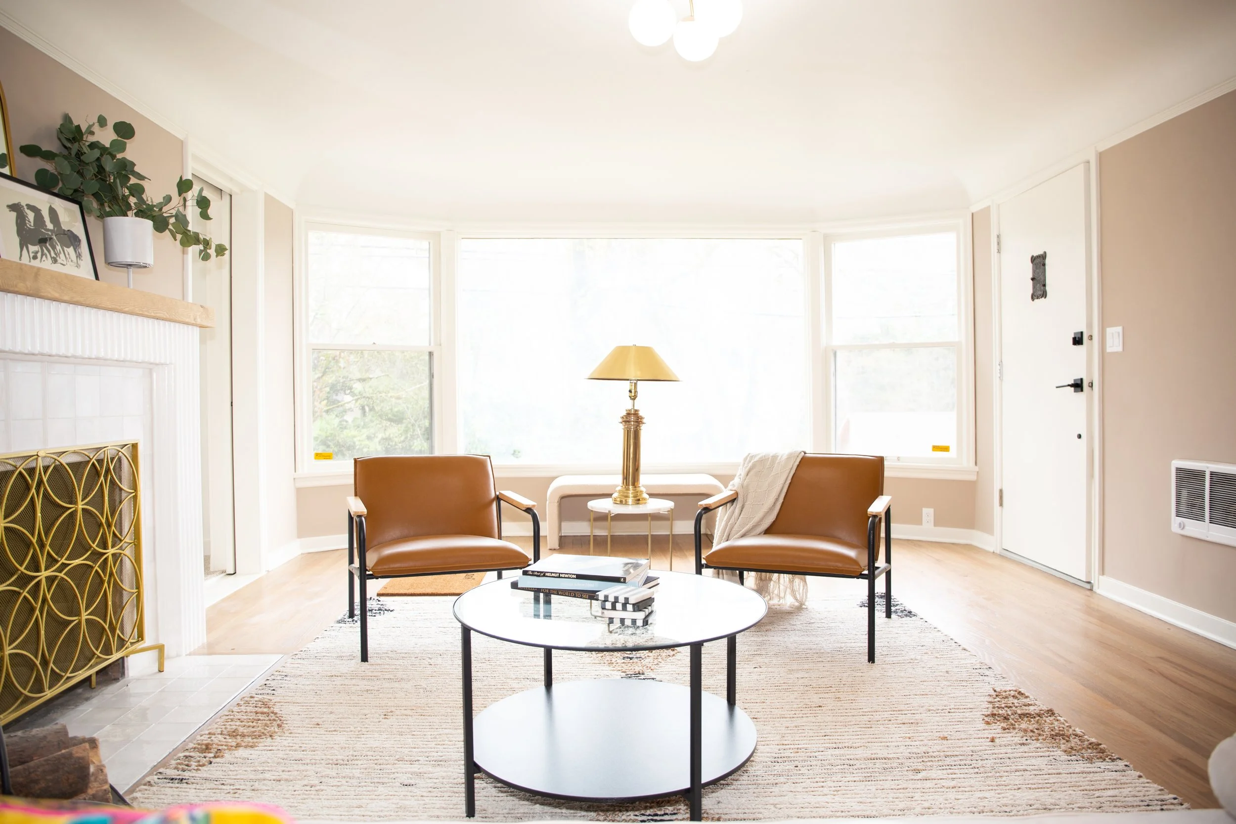 Bright living room with large bay windows, two brown leather armchairs, a round black coffee table with books, a gold table lamp, patterned area rug, and a white fireplace with decorative items and greenery on the mantel.