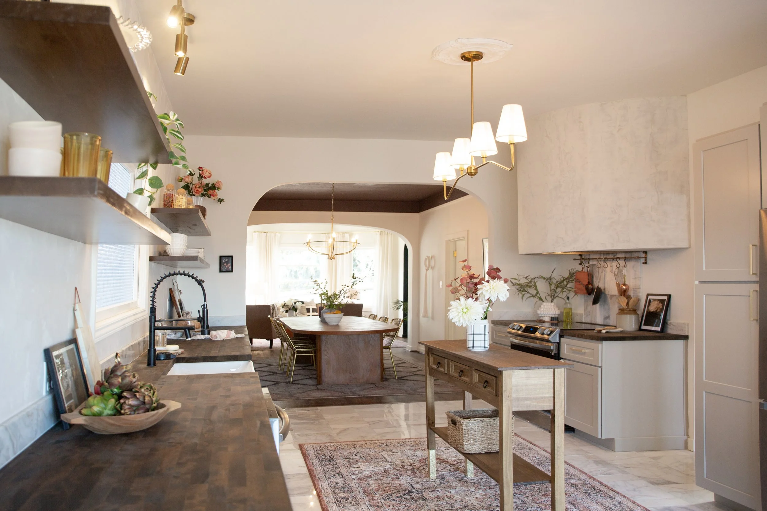 A spacious and well-lit kitchen with wooden countertops, open shelves, a small central table with storage, and a view into a dining and living area with large windows and floral decorations.