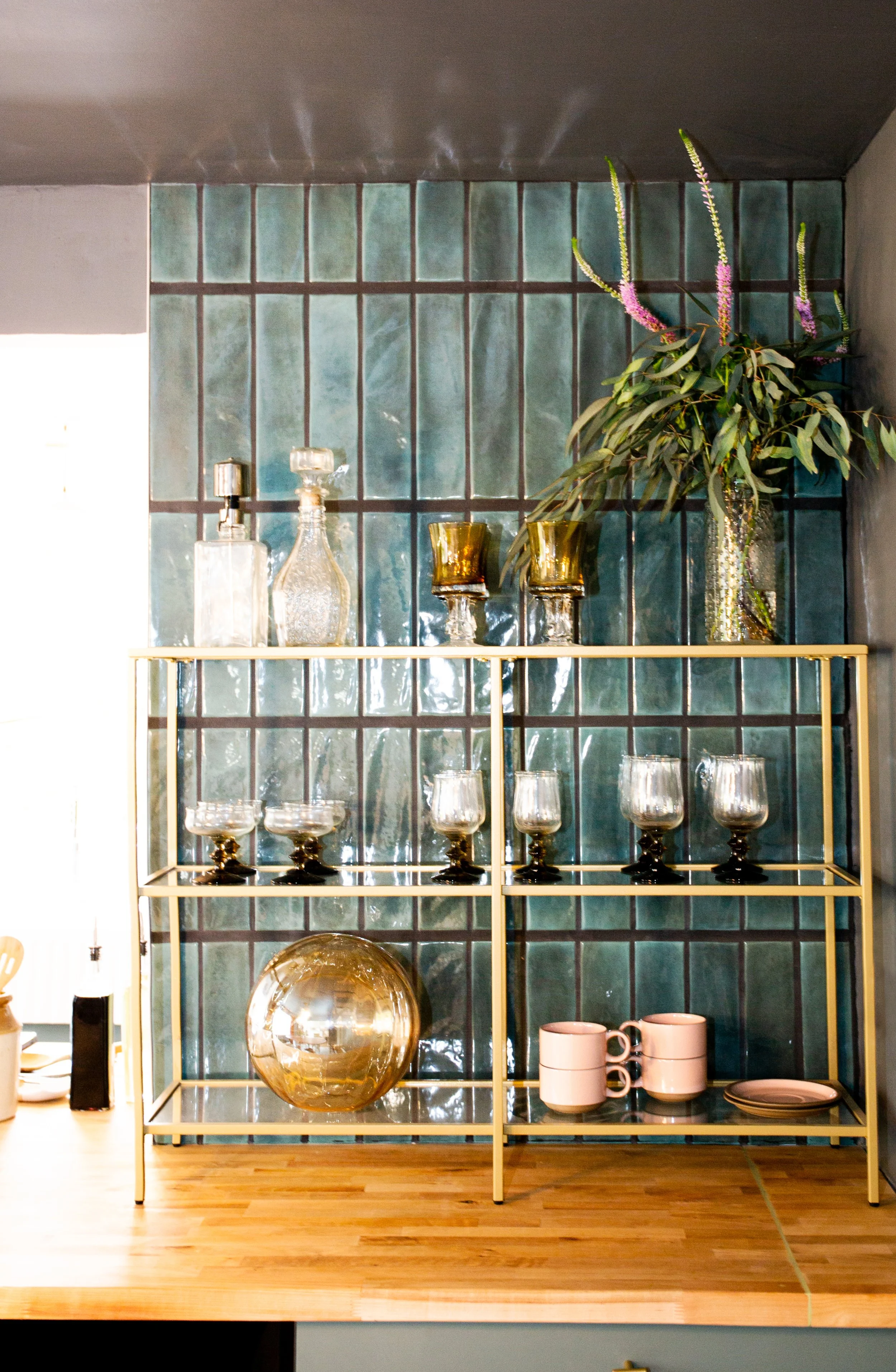 Decorative glassware and a vase with greenery on a gold shelving unit against a tiled wall in a kitchen or dining area.